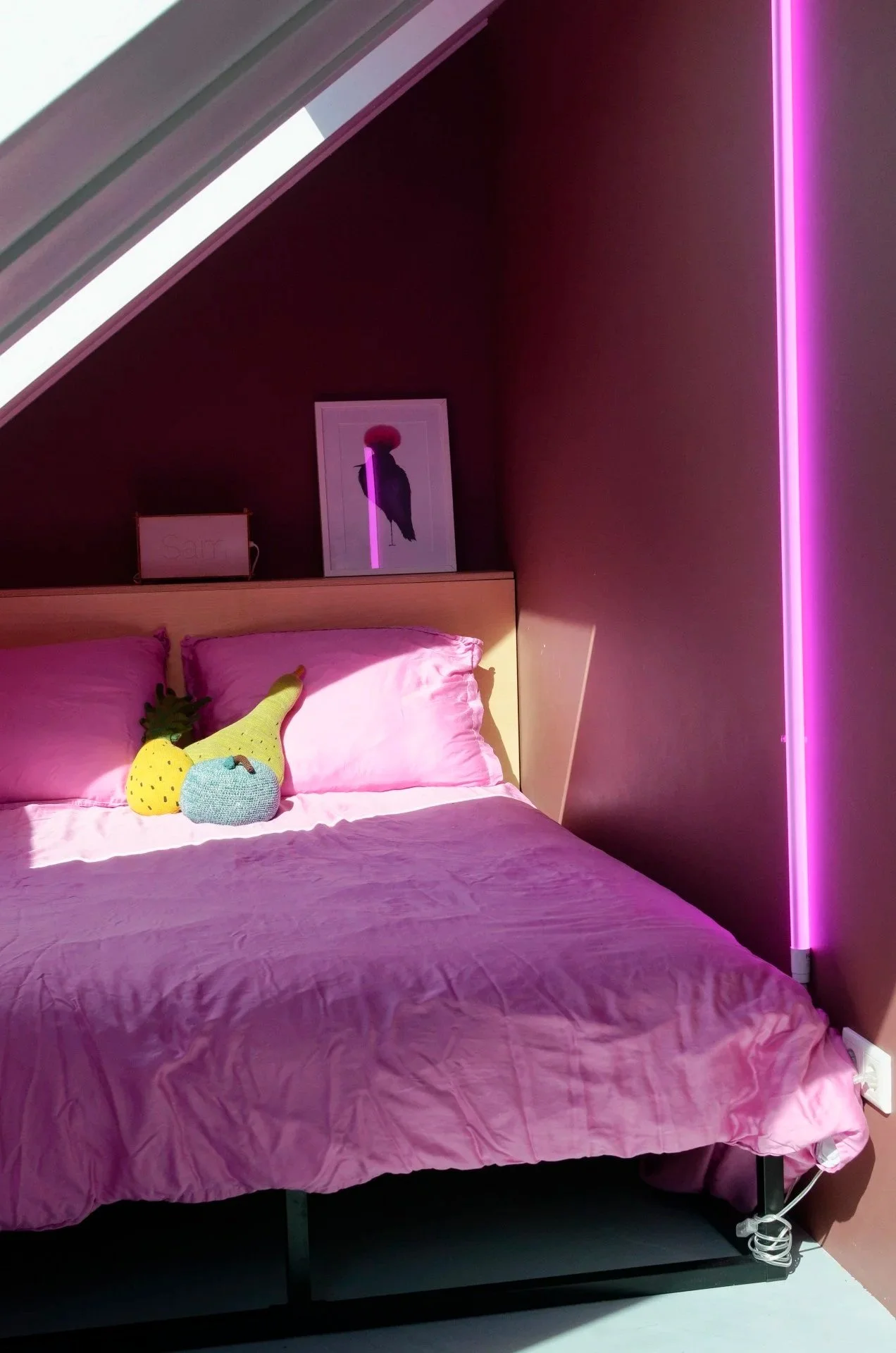 A bed with pink bedding and a knitted pineapple-shaped pillow in a room with dark walls and pink neon lighting.