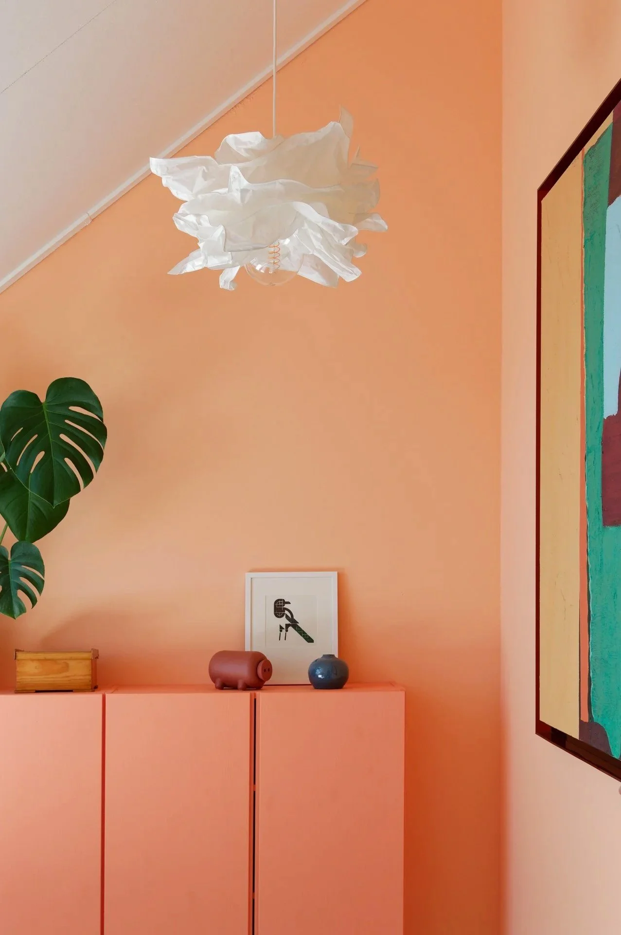 Interior of a room with salmon-colored walls, a modern white paper lantern pendant light, a large green monstera plant, framed artwork with abstract design, a giant pink pig-shaped object, and two small round vases on a salmon-colored cabinet.