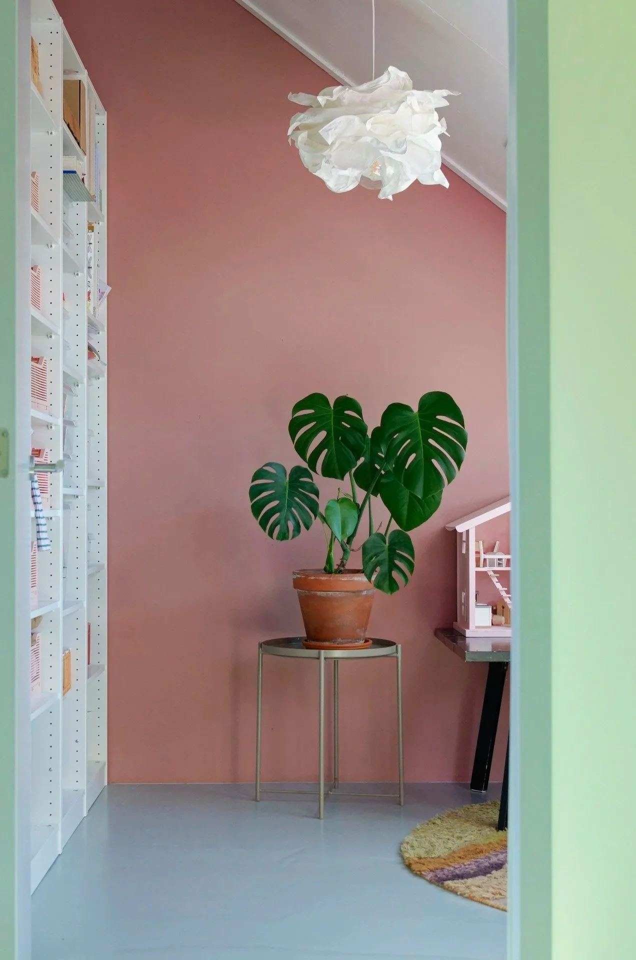 A view through a doorway into a room with a pink wall, white ceiling, and a pink and white pendant light fixture. There is a large potted Monstera plant on a small round table and part of a black table with a miniature dollhouse in the corner. A circular rug with multicolored stripes is on the floor.