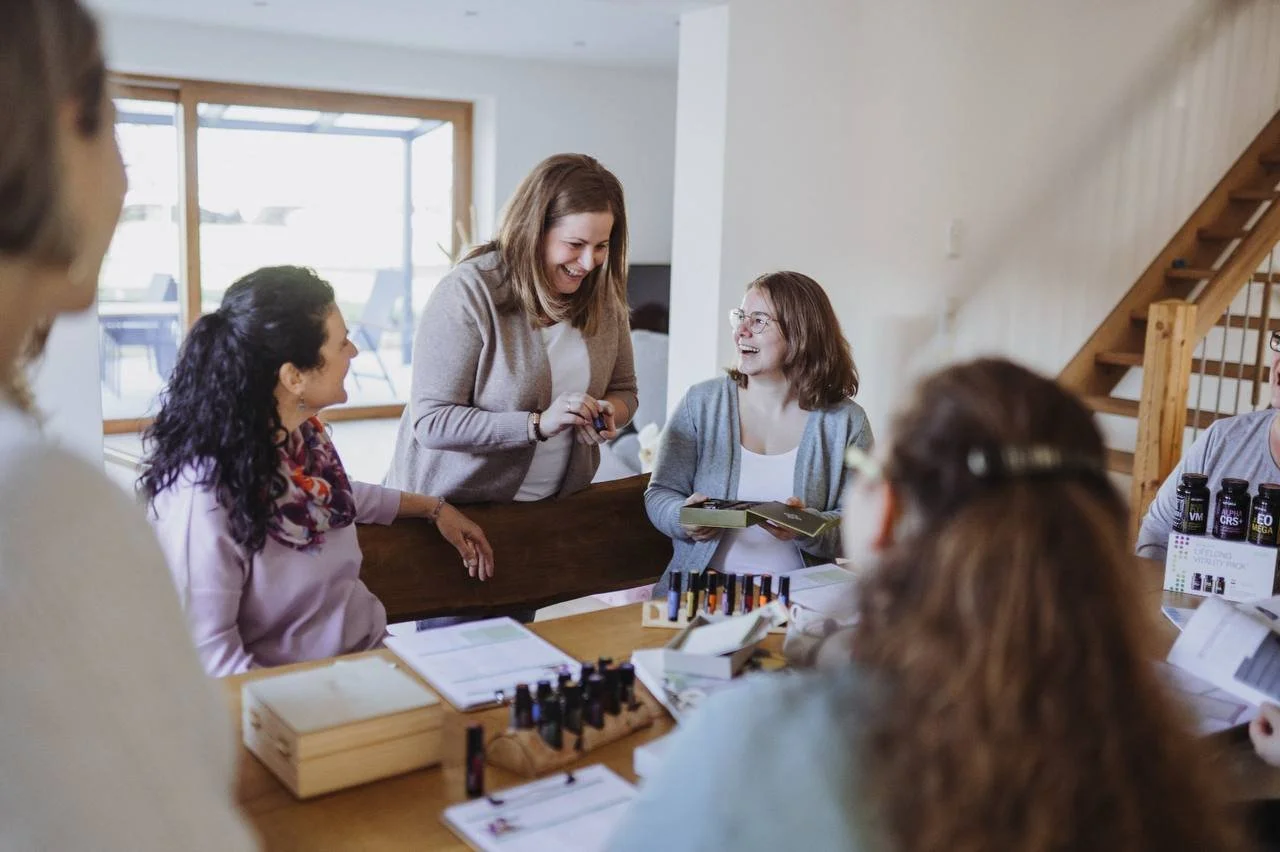 Gruppe von Frauen bei einem Treffen, beim Aromatherapie-Workshop, in einem Raum mit großen Fenstern und Holztreppe, lachend und bei der Produktpräsentation.