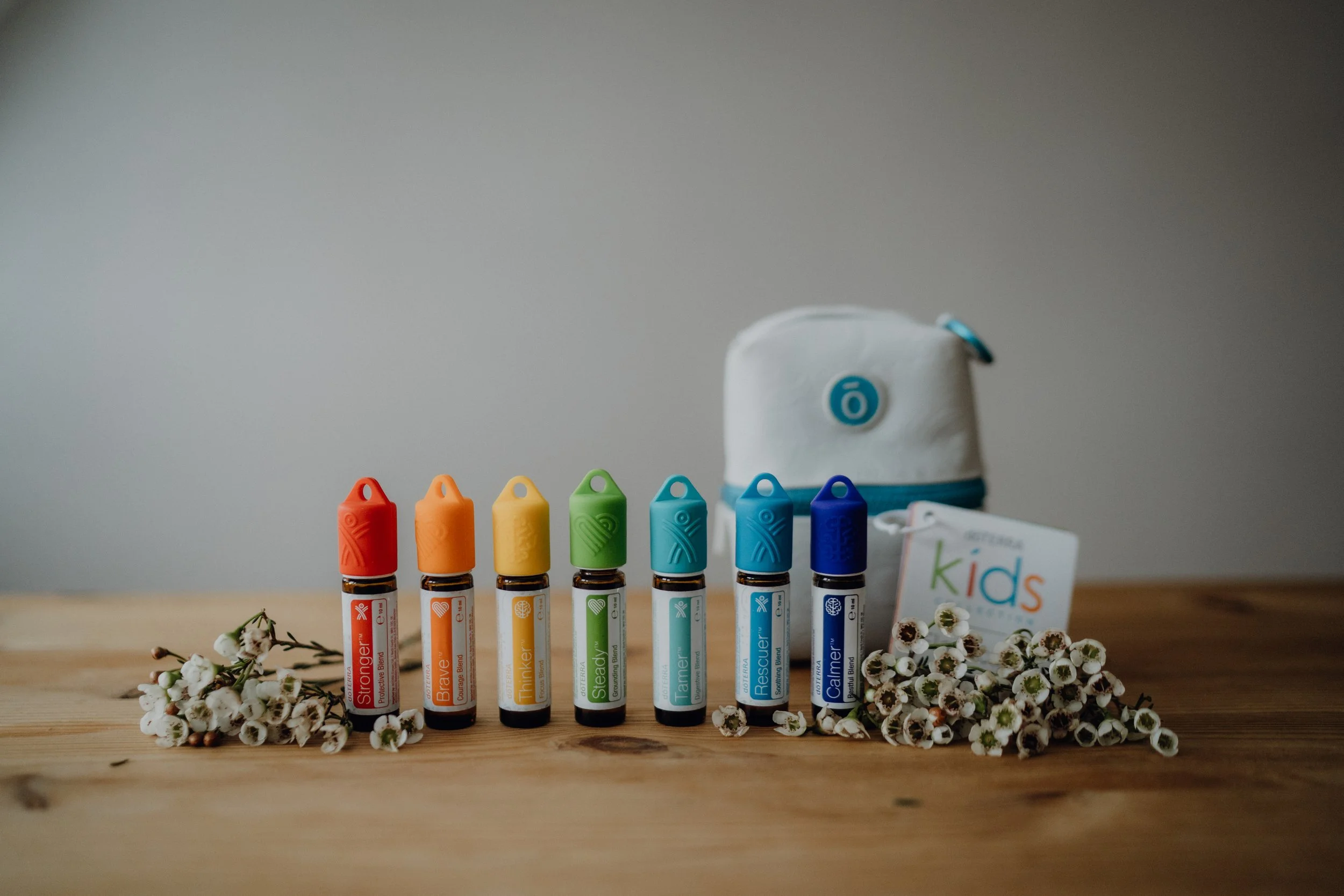 Sieben ätherische Ölroller für Kinder auf einem Holztisch, die verschiedenen Farben und Beschriftungen haben, um Kinder beim Lernen zu unterstützen, mit weißen Blumen und einem weißen Beutel im Hintergrund.