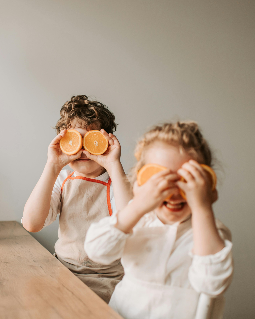 Zwei lachende Kinder spielen mit halben Orangen vor ihren Augen, in einem hellen Raum an einem Holztisch.