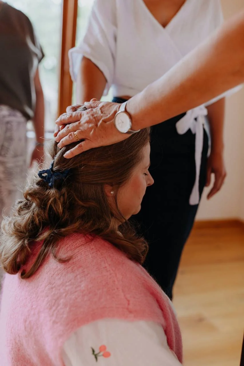 Eine Frau mit geschlossenen Augen erhält eine Handauflage auf den Kopf, während eine andere Person sie sanft berührt. Es wirkt wie eine spirituelle oder heilende Zeremonie als Teil einer ganzheitlichen Familienaufstellung.