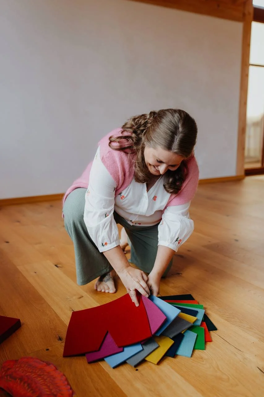 Eine Familienaufstellerin sortiert farbige Bodenanker aus Filz für ganzheitliche Familienaufstellungen auf Holzfußboden. Sie trägt eine weiße Bluse mit Blumenmuster und eine pinkfarbige Strickjacke.