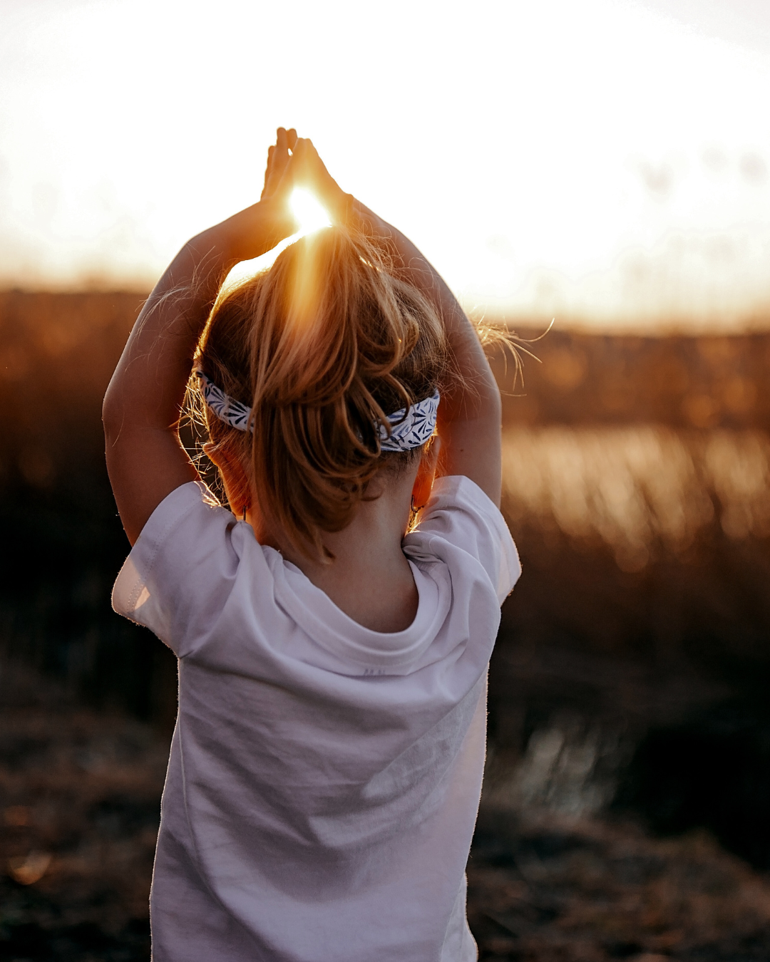 Ein junges Mädchen steht draußen bei Sonnenuntergang, mit den Händen über dem Kopf und scheint den Sonnenstrahl zu begrüßen oder zu berühren.