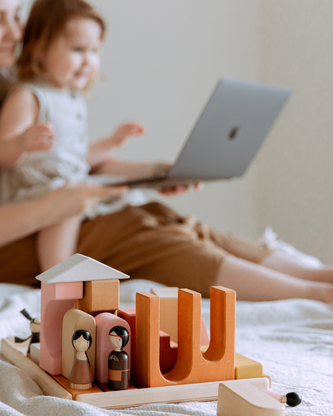 Ein Spielzeughaus mit zwei Figuren und Holzklötzen auf einem Bett, mit einer verschwommenen Frau und Kind im Hintergrund, die an einem Laptop arbeitet.