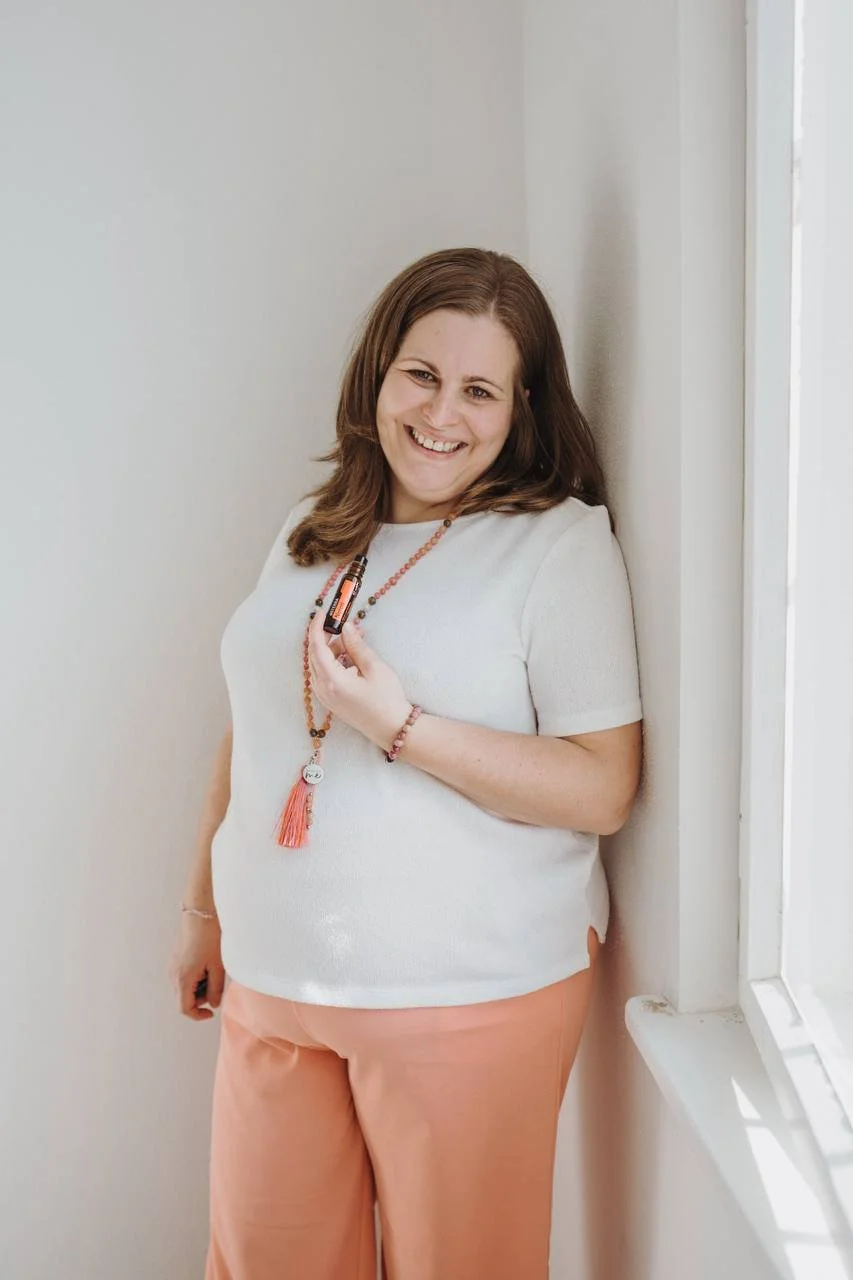 Eine lächelnde Frau lehnt an einer Wand neben einem Fenster, trägt ein weißes Oberteil und peachfarbene Hosen, hält ätherische Öle in der Hand, trägt eine Edelsteinkette und ist gut gelaunt.