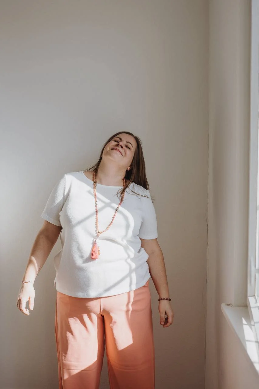 Eine lachende Frau steht in einem Raum neben einem Fenster, das Sonnenlicht hineinscheinen lässt.