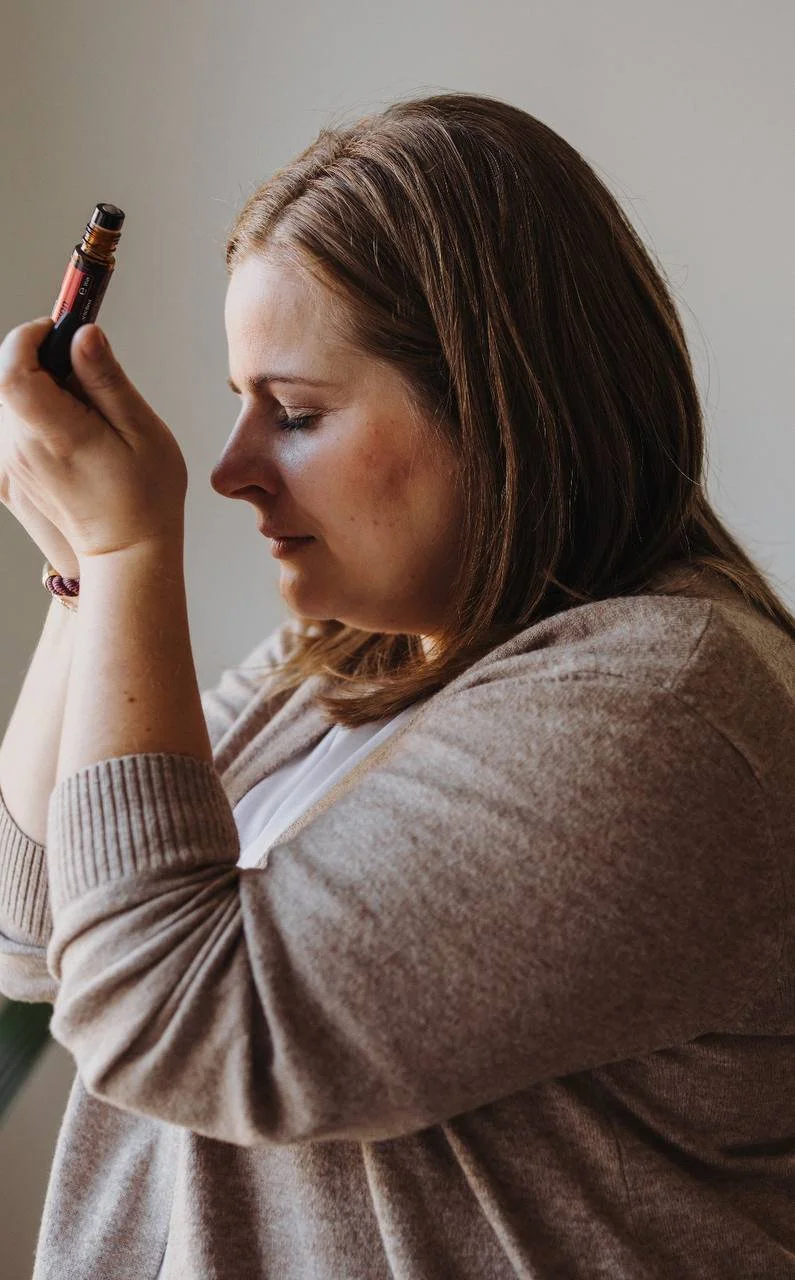 Eine Frau mit braunen Haaren inhaliert mit einem tiefen bewussten Atemzug den Duft eines ätherischen Öls mit geschlossenen Augen. Sie trägt eine beigefarbene Jacke.