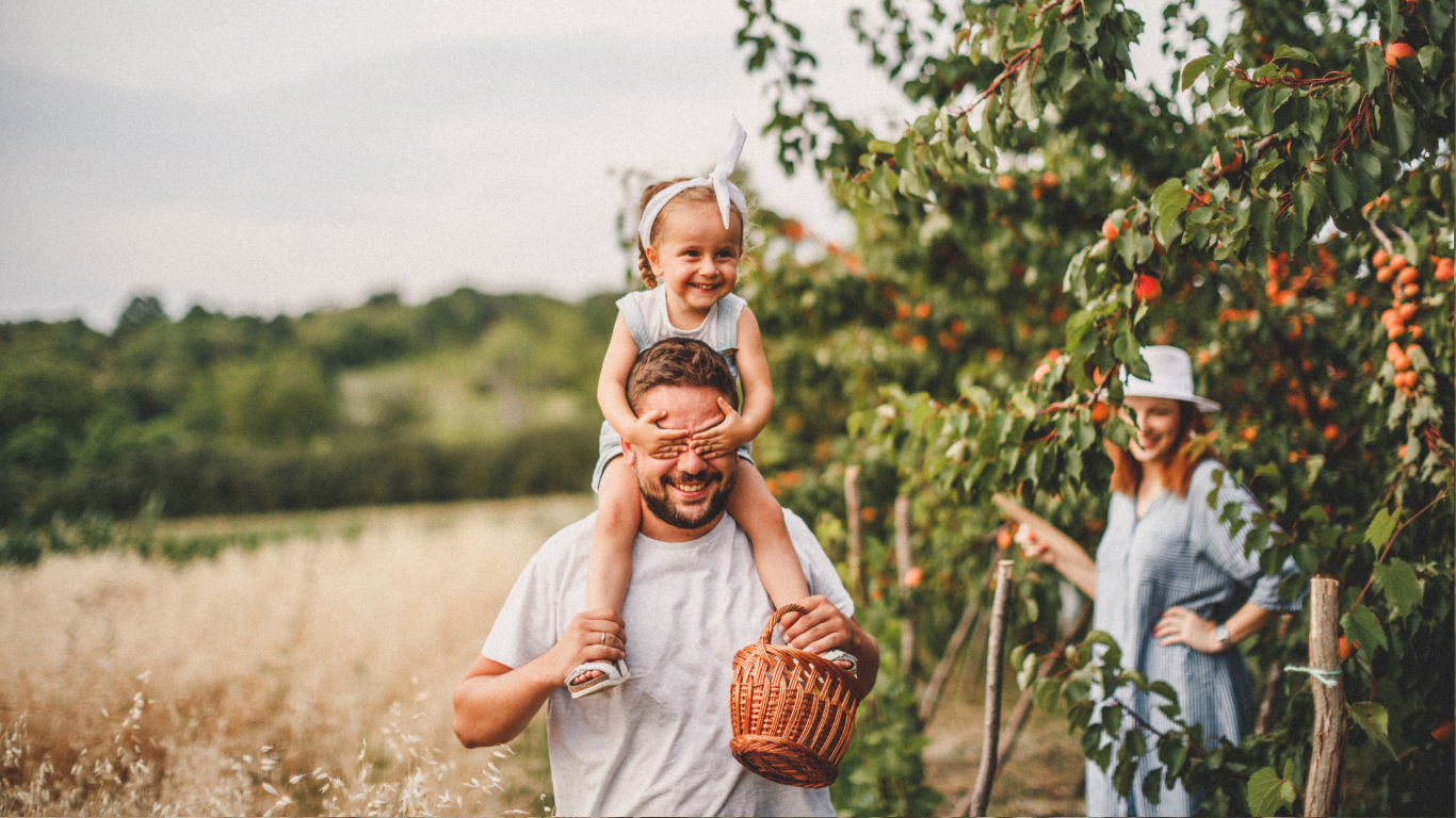 Ein Mann trägt ein lachendes Mädchen auf den Schultern durch eine Apfelplantage, während eine Frau im Hintergrund eine Hand auf eine Baumreihe legt.