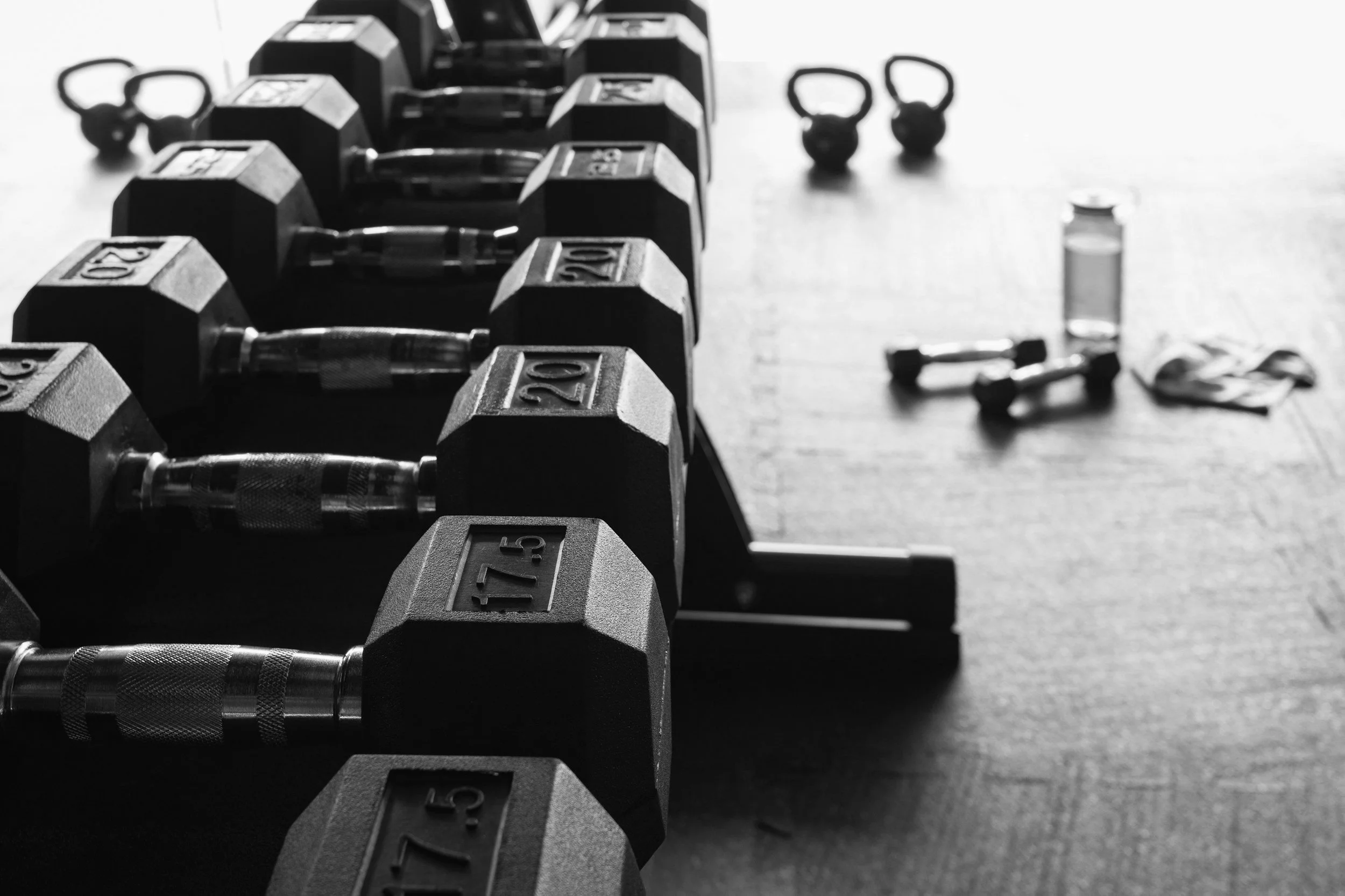 A row of dumbbells with weight labels ranging from 15 to 20 pounds lined up on a workout floor. In the background, there are kettlebells, a water bottle, and a towel on the floor.