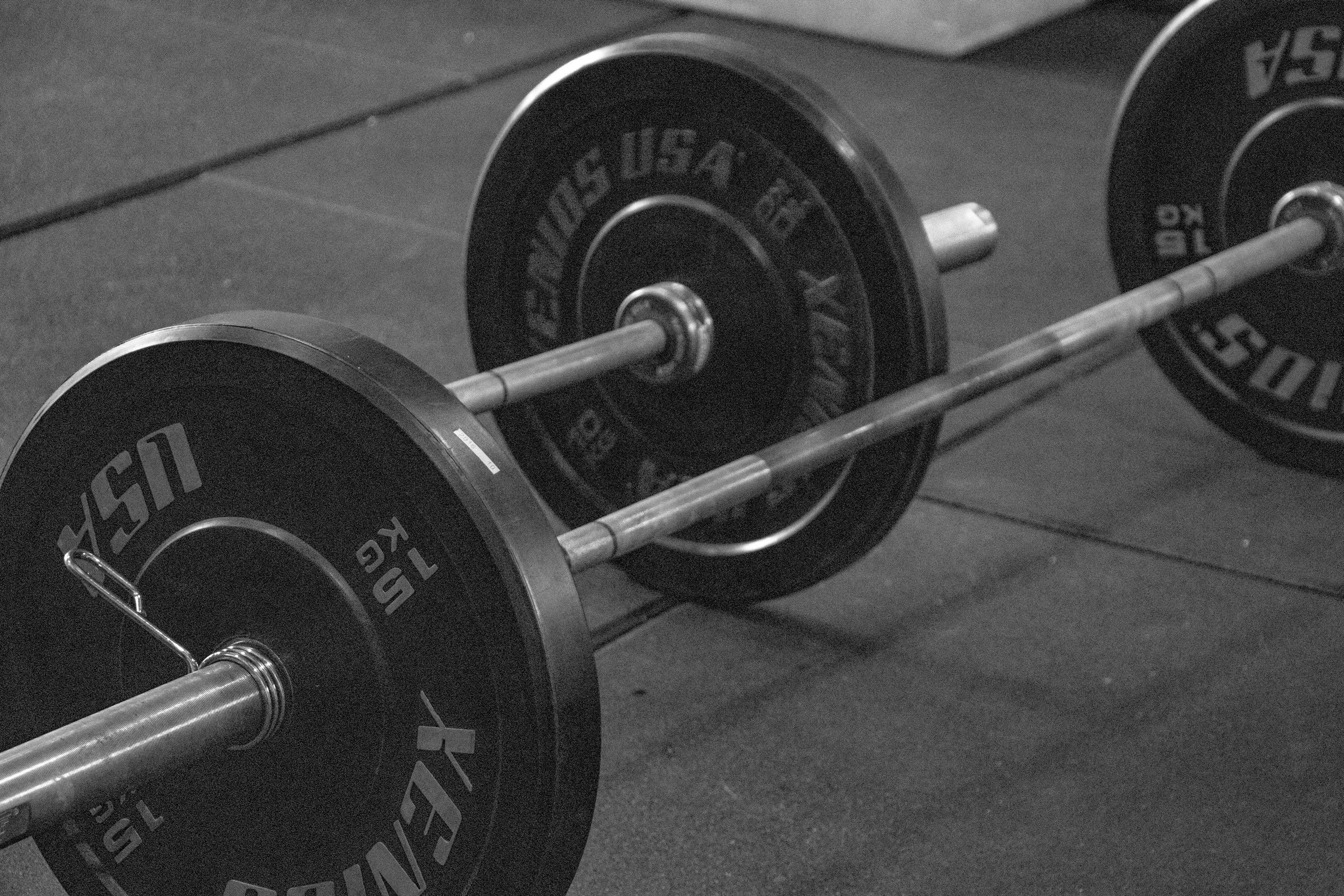 A black and white photo of a barbell with weights on a gym floor.