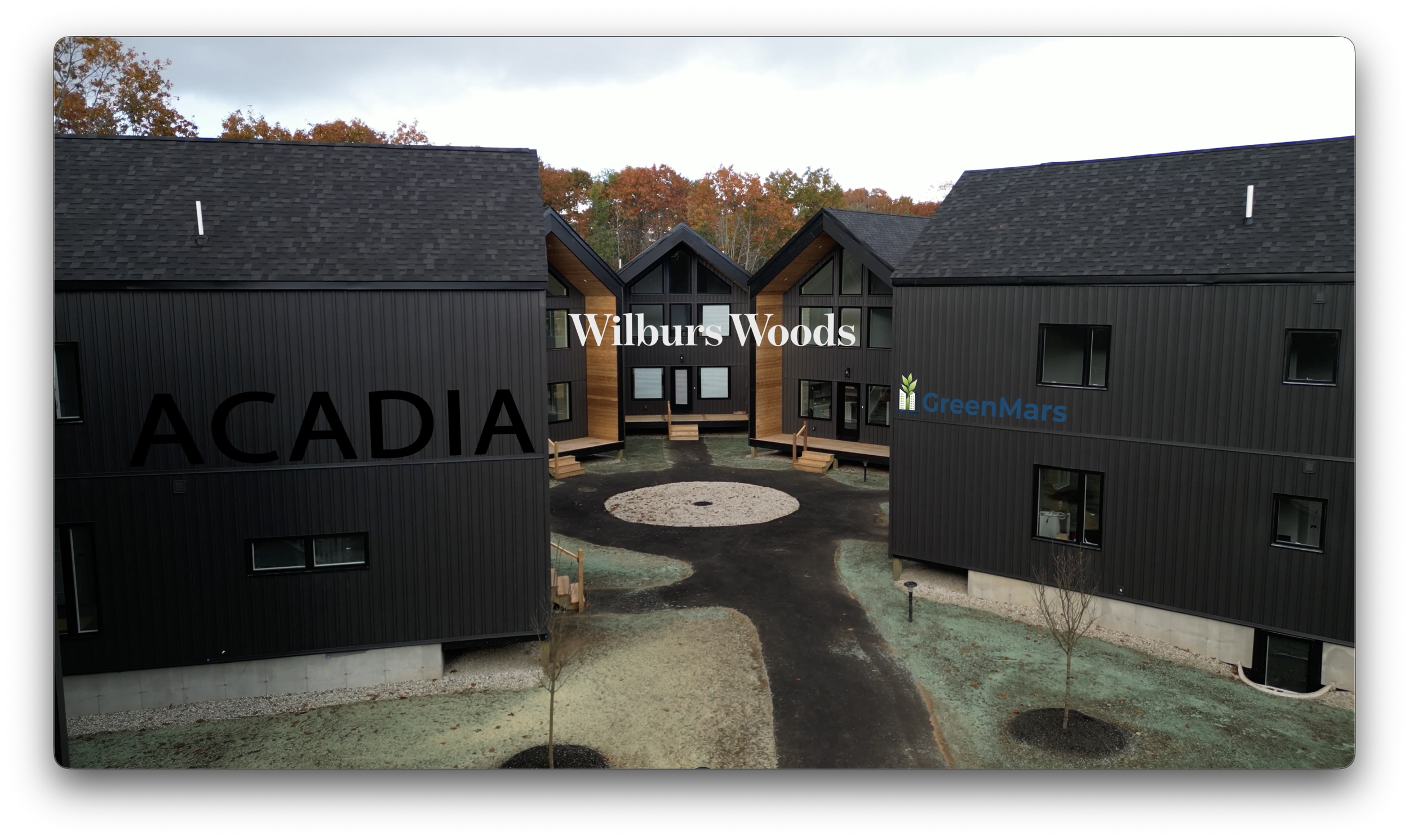 Modern black residential buildings with gabled roofs, with the names Wilbur Woods, ACADIA, and GreenMars labeled on their facades, surrounded by a landscaped courtyard with small trees and pathways.