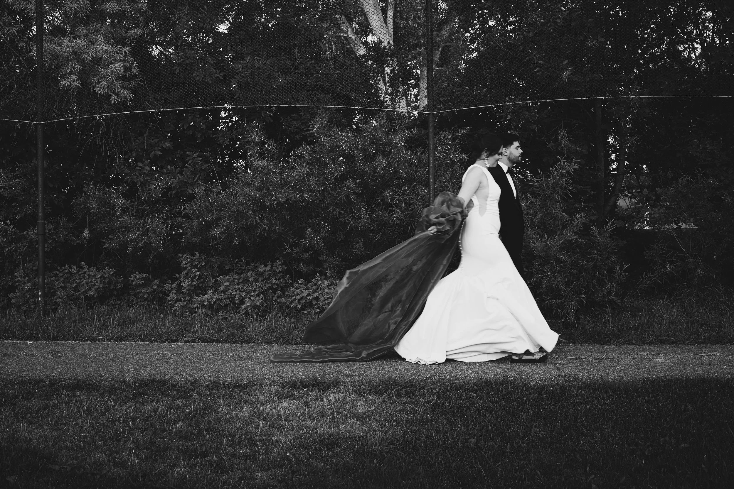 A black and white photo of a bride and groom walking outside on a sidewalk, holding hands, with a dark, leafy background.