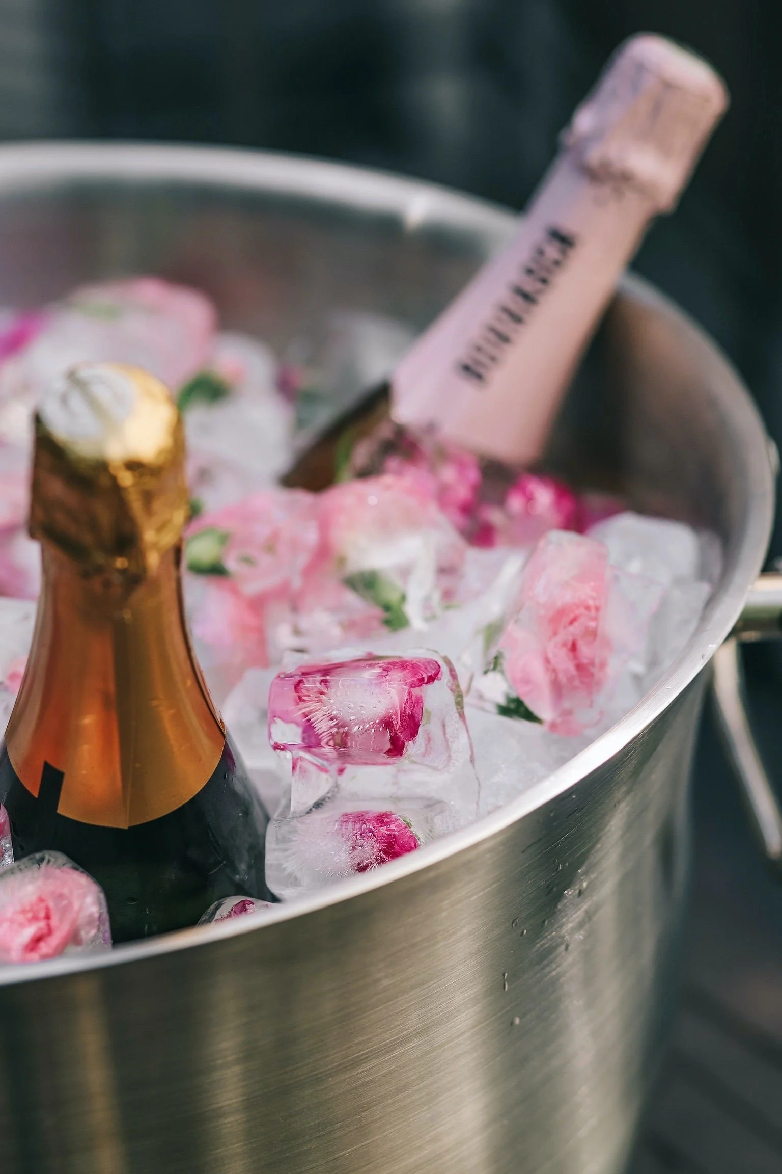 A stainless steel ice bucket filled with ice, pink roses, a bottle of champagne, and a bottle of rosé wine.