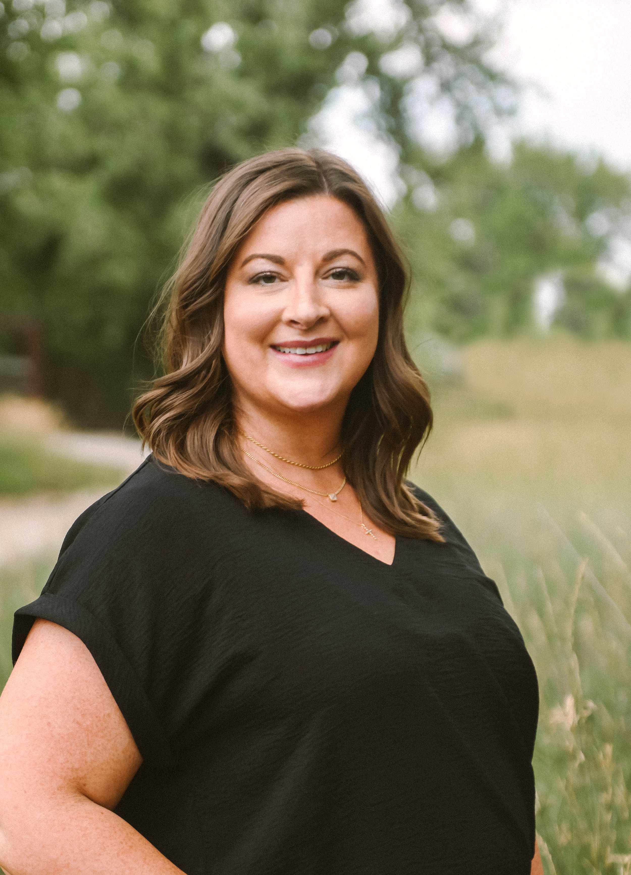 A woman with shoulder-length brown hair wearing a black top and layered gold necklaces, smiling outdoors with green trees in the background.