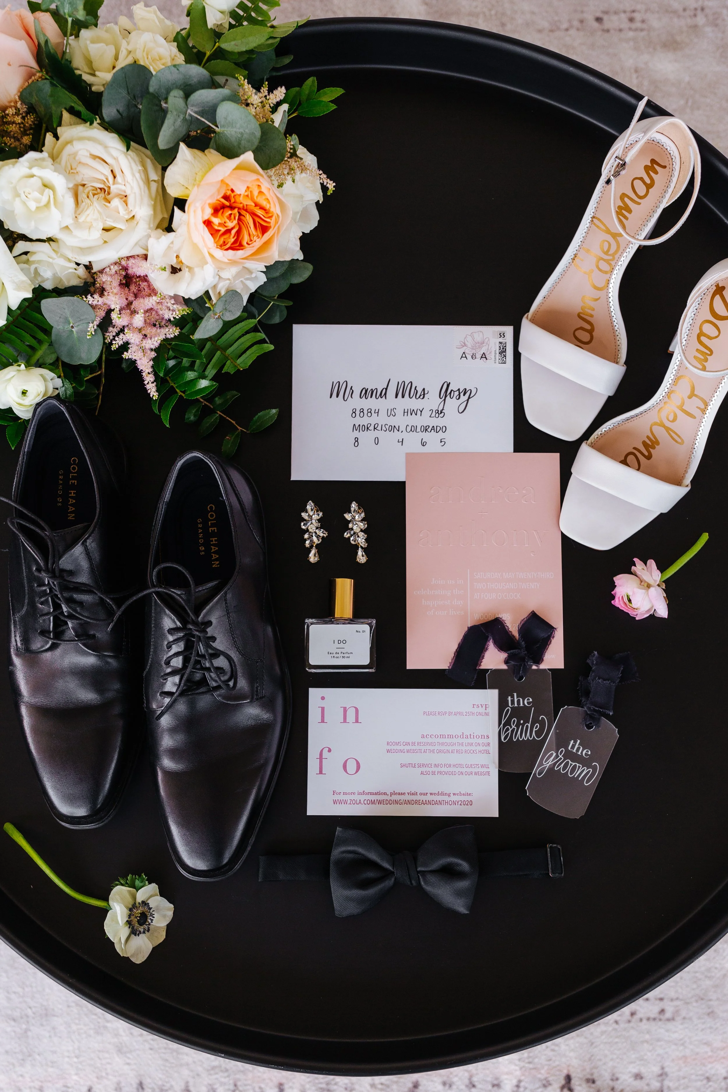 Wedding accessories and invitation on a black round table, including a bouquet of white and peach roses, white high-heeled shoes with gold lettering, black dress shoes, a pair of earrings, a perfume bottle, a black bow tie, and wedding invitations and tags for bride and groom.