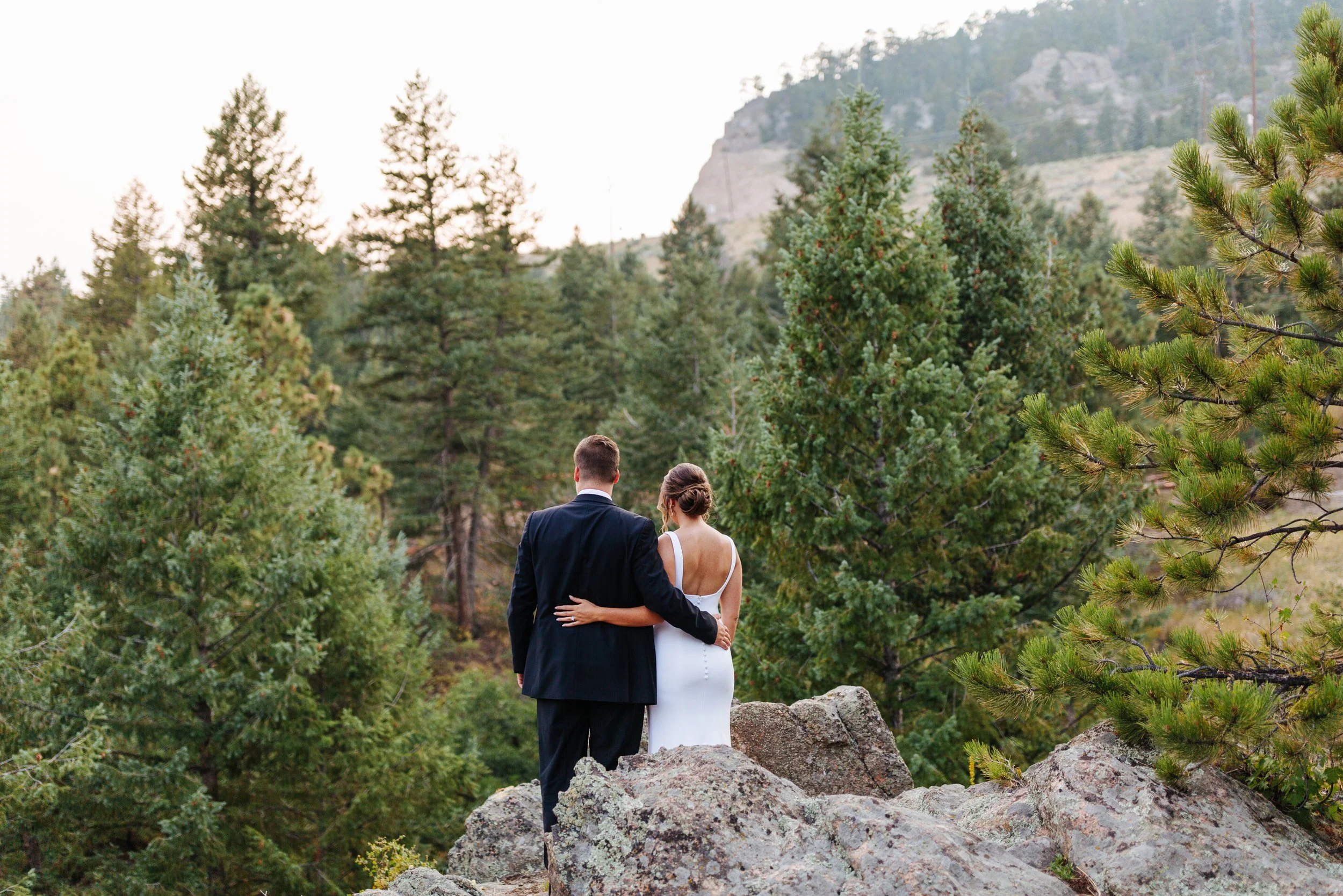 A couple in wedding attire standing on rocks in a forested area with green pine trees and mountains in the background.
