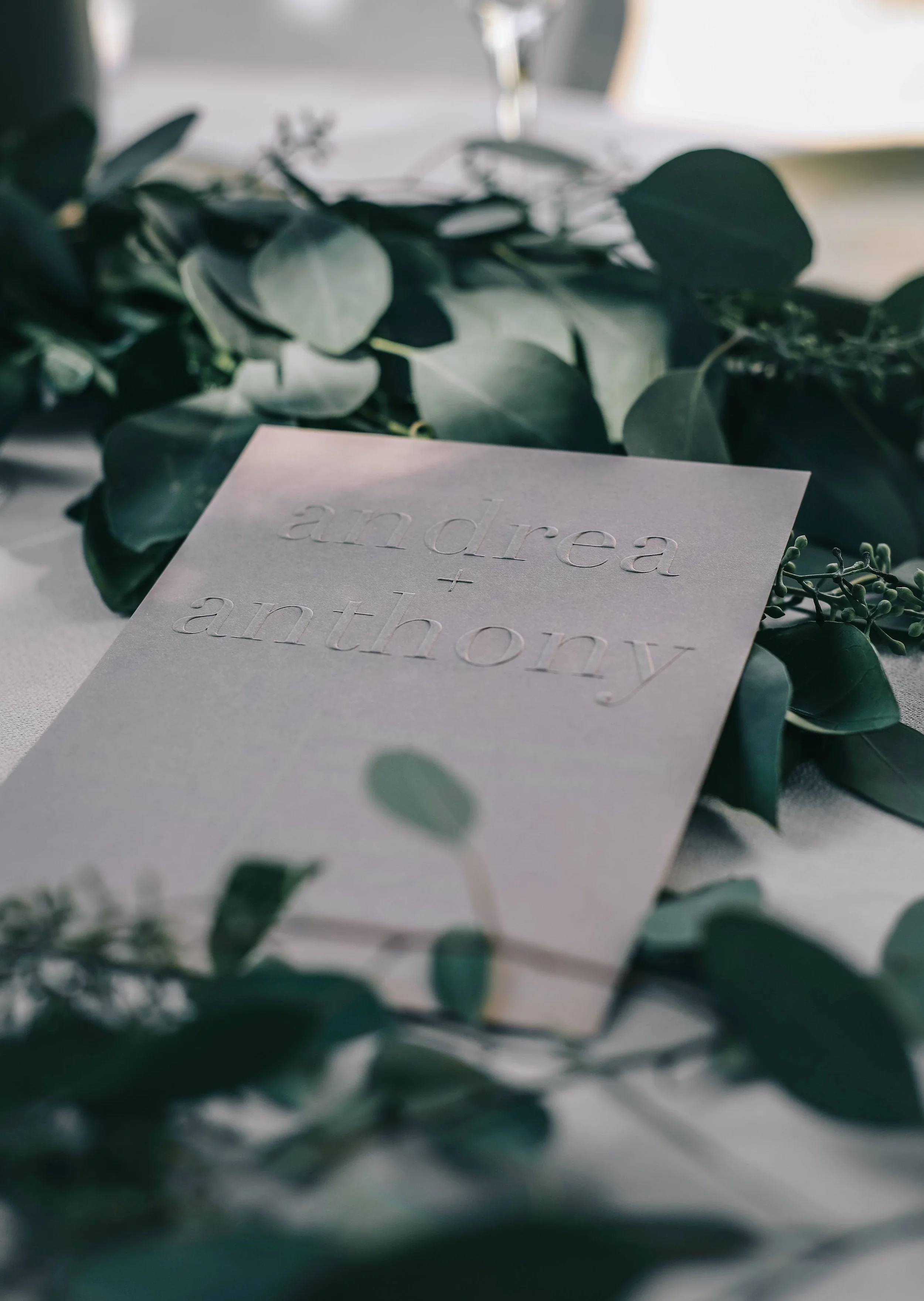 Wedding invitation with embossed text 'andrea + anthony' surrounded by greenery.