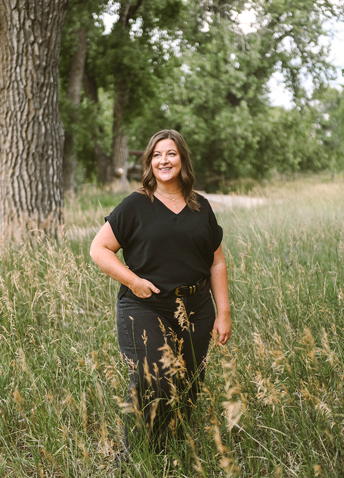 A woman with shoulder-length brown hair, wearing a black V-neck top and black pants, stands in a grassy field near large trees, smiling and looking off to the side.