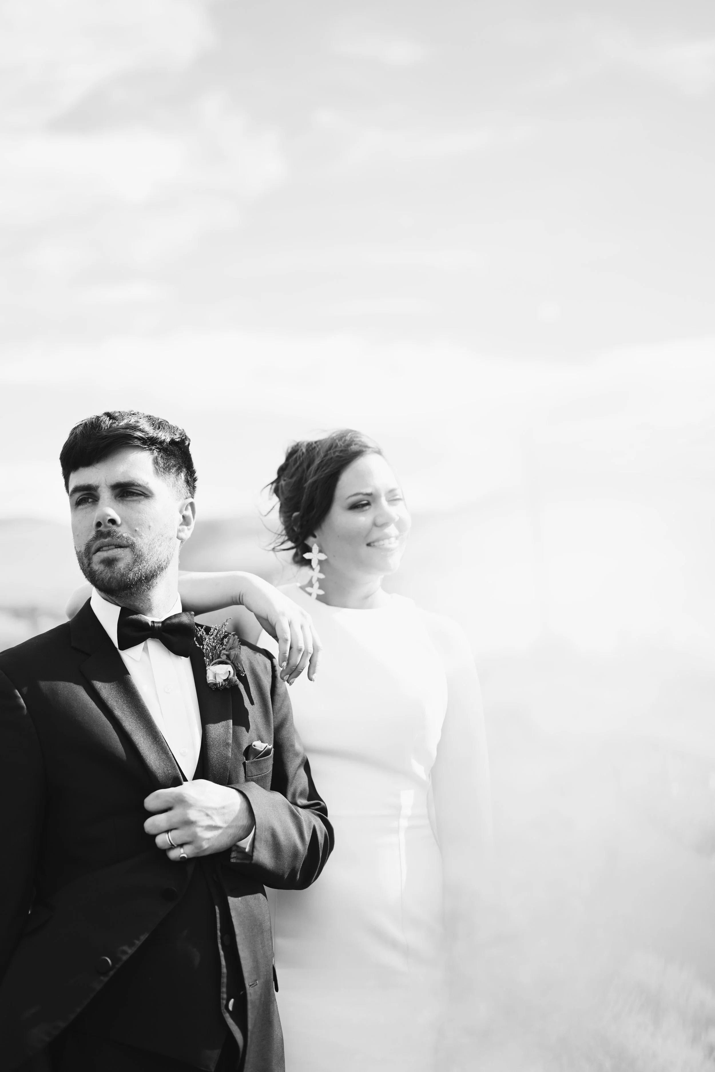 Black and white photo of a groom in a tuxedo and a smiling bride in a white dress, outdoors on a bright day.