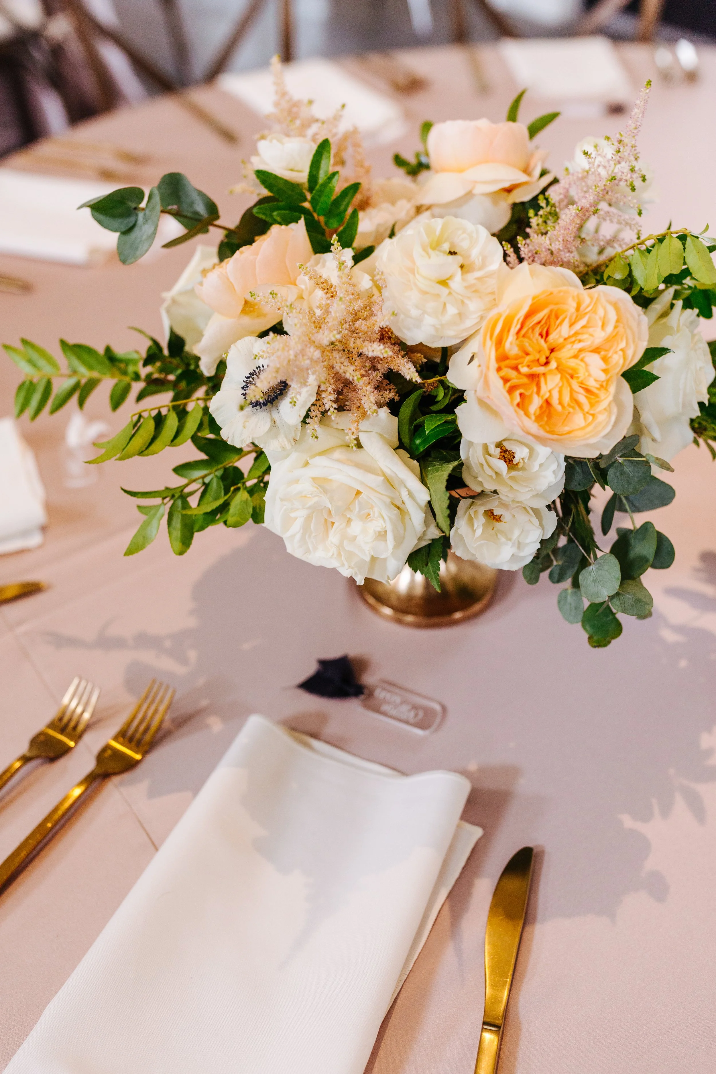 A floral centerpiece with white and peach roses and greenery on a table for a formal event.