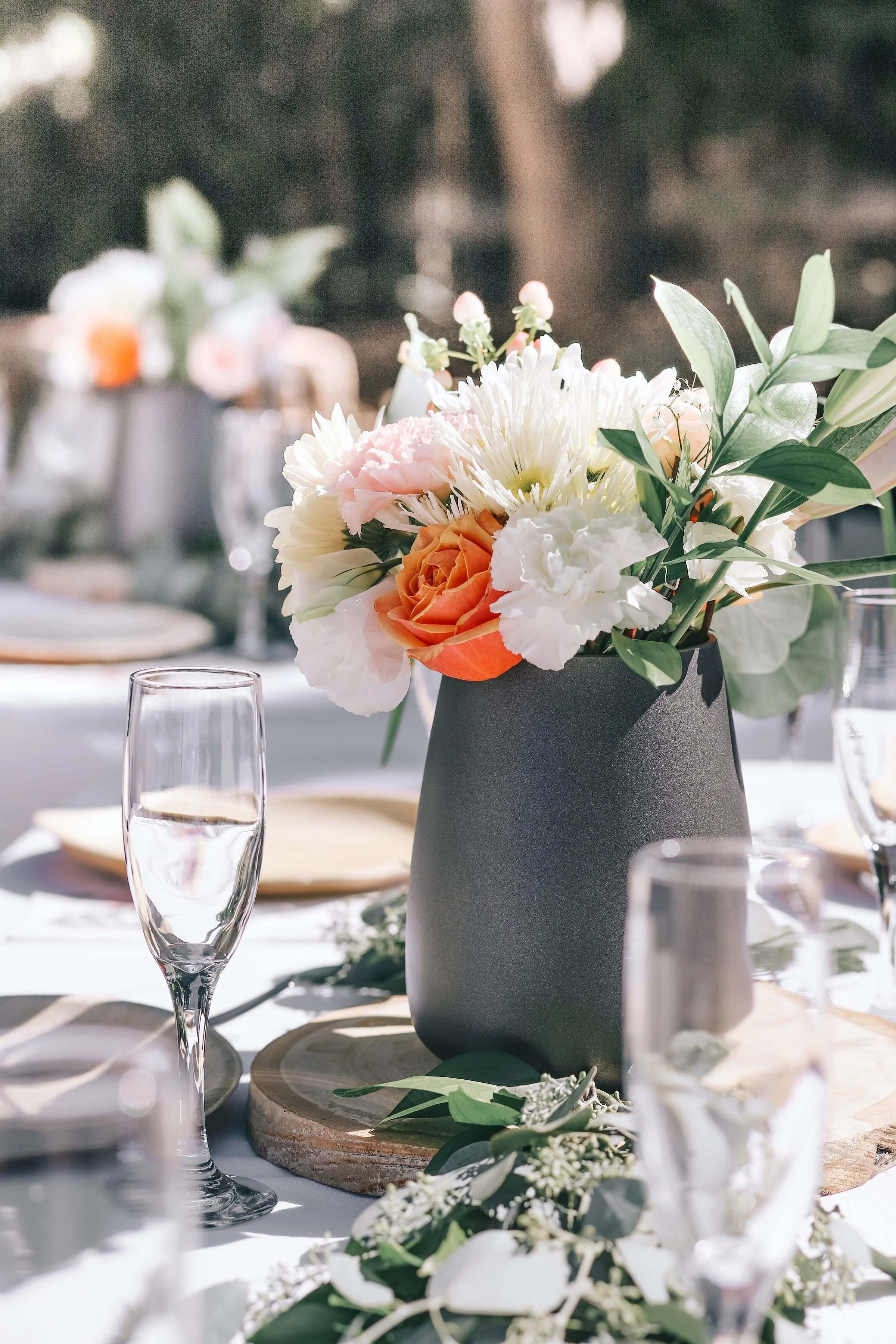 A floral centerpiece with white, pink, and orange flowers in a black vase on a table at an outdoor event.