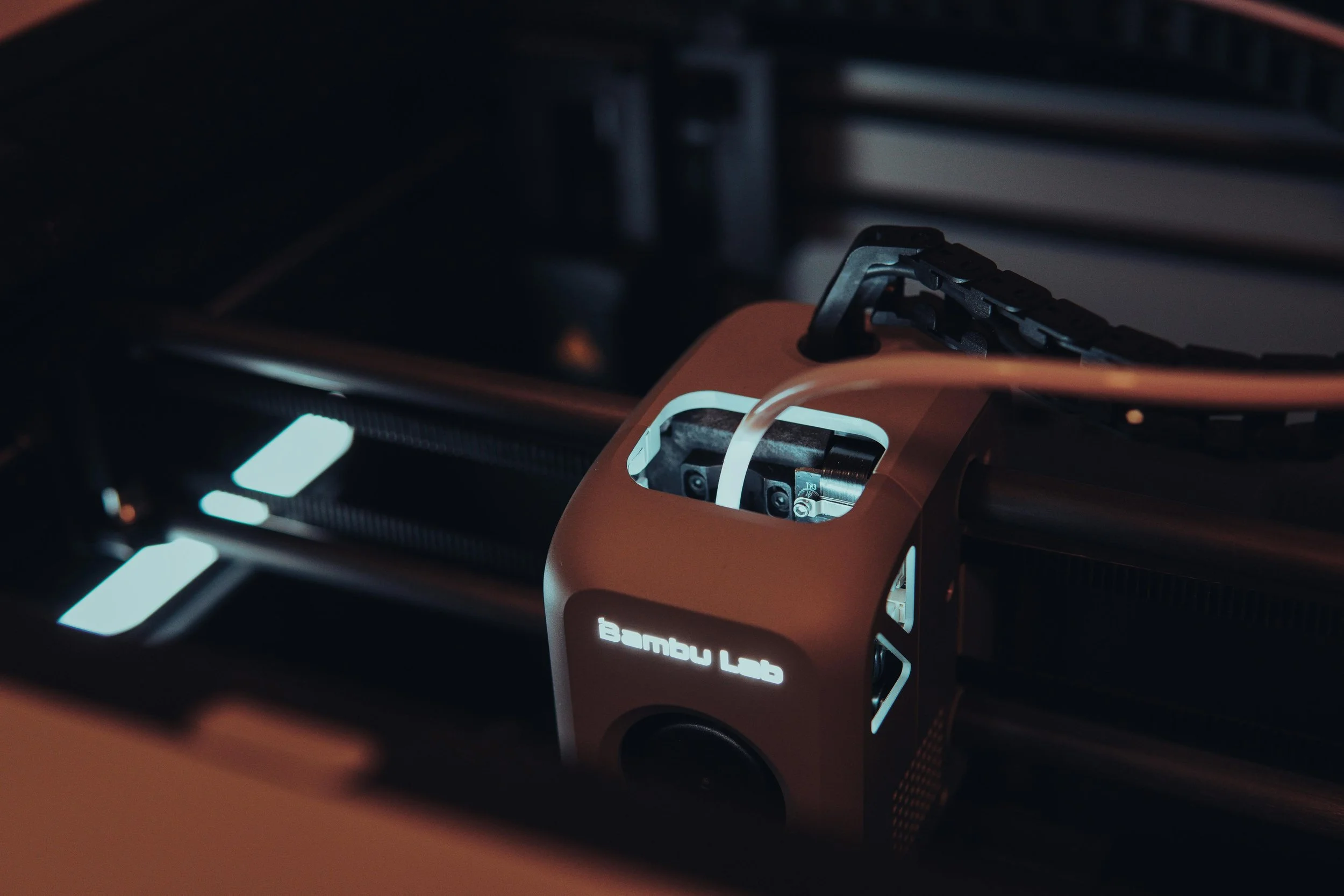 Close-up of a Bambu Labs 3D printer extruder and hotend with the logo illuminated, showing the internal components and filament path.