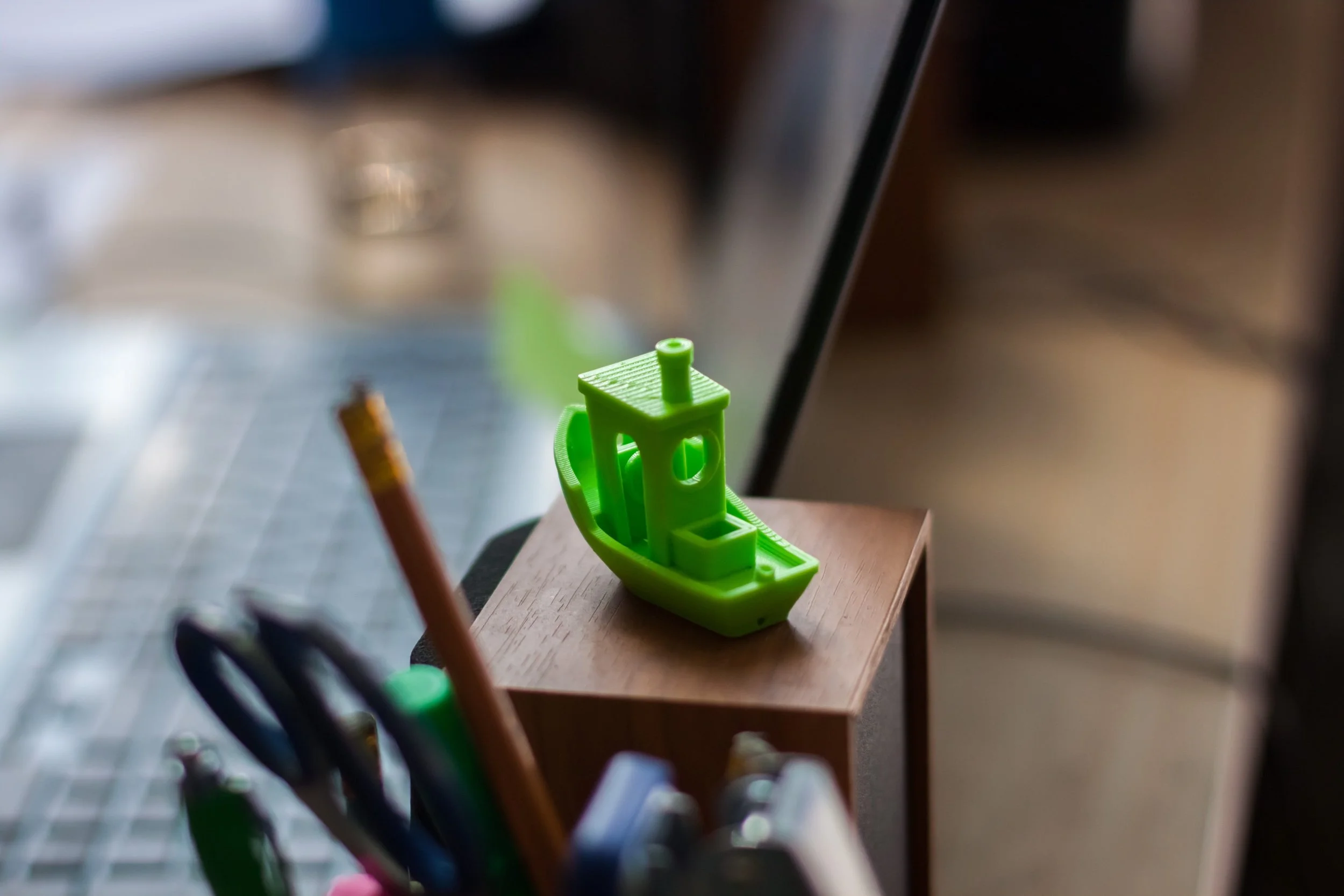 A small 3D printed toy boat on a wooden surface, with office supplies in the foreground and a blurred background.