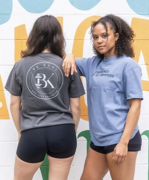 Two young women standing in front of a colorful mural, one with her back facing the camera and the other facing forward, both wearing casual T-shirts and shorts.