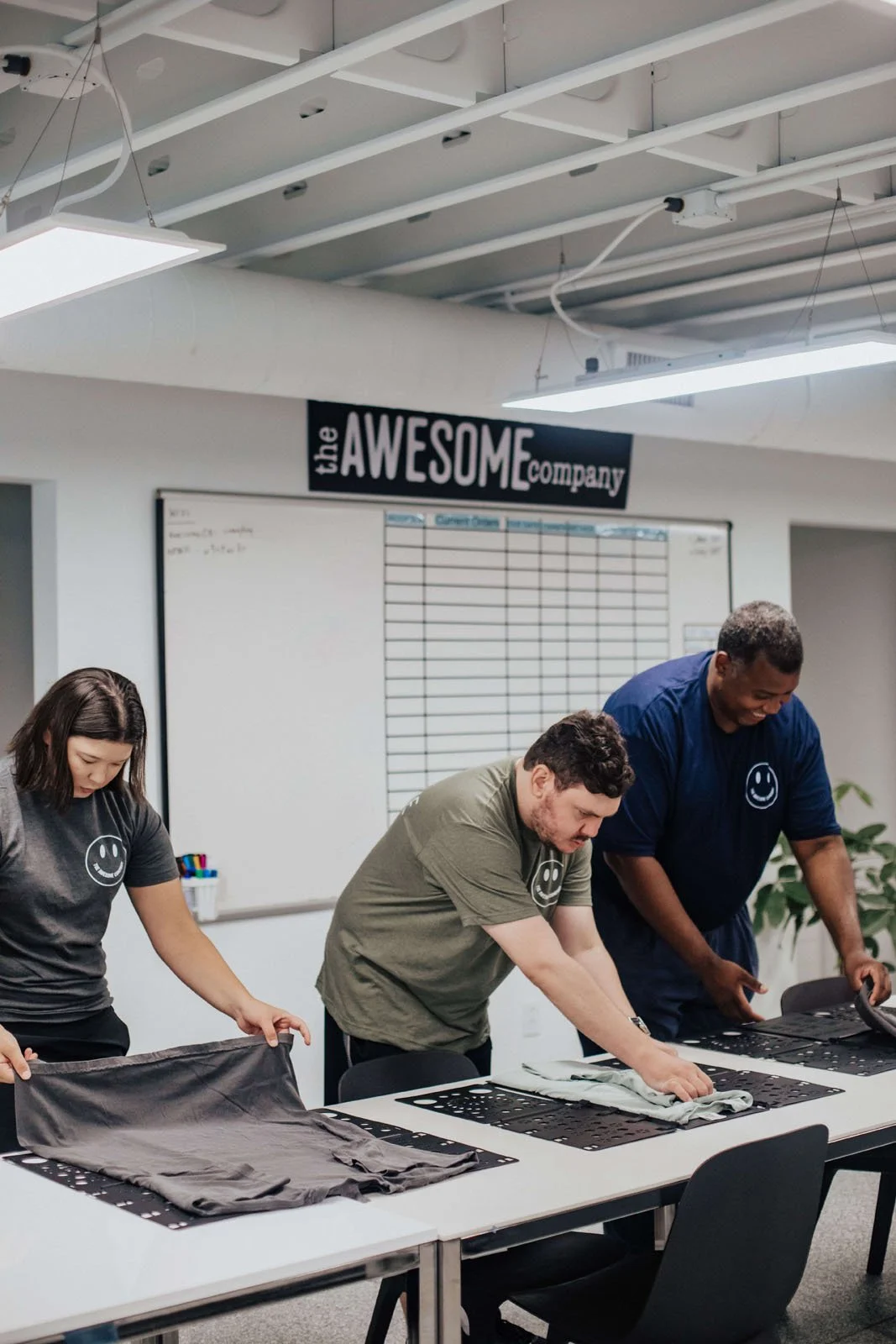 Three people folding T-shirts at a table in a meeting room with whiteboard and sign reading 'the AWESOME company'