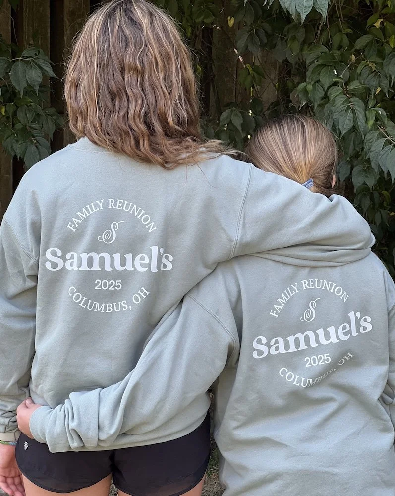 Two people wearing matching gray sweatshirts with a family reunion message, standing outdoors with green leaves in the background.