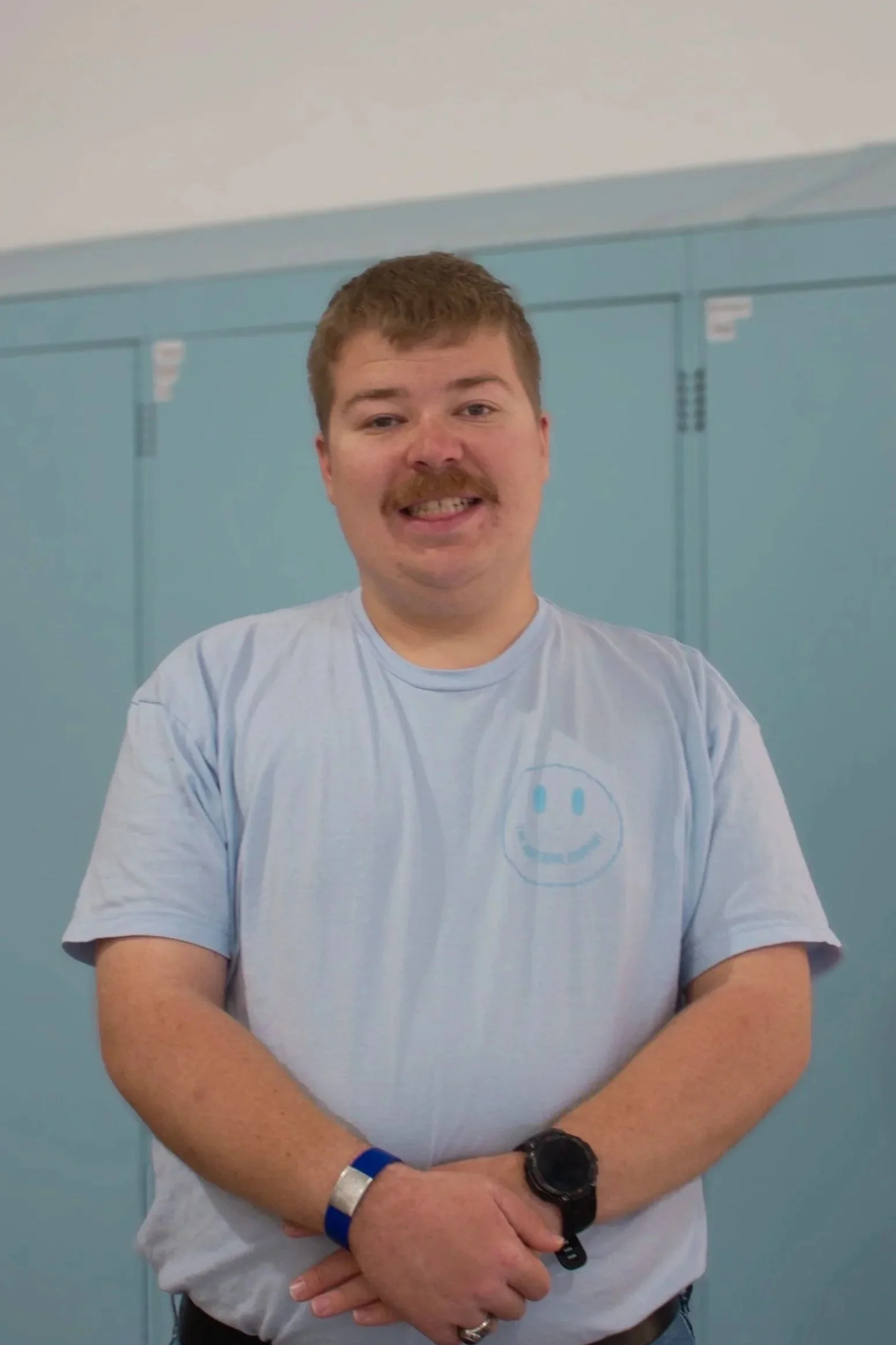 A young man with light hair, and a mustache beard, wearing a green T-shirt with a smiley face logo and black pants, standing in front of light blue lockers.