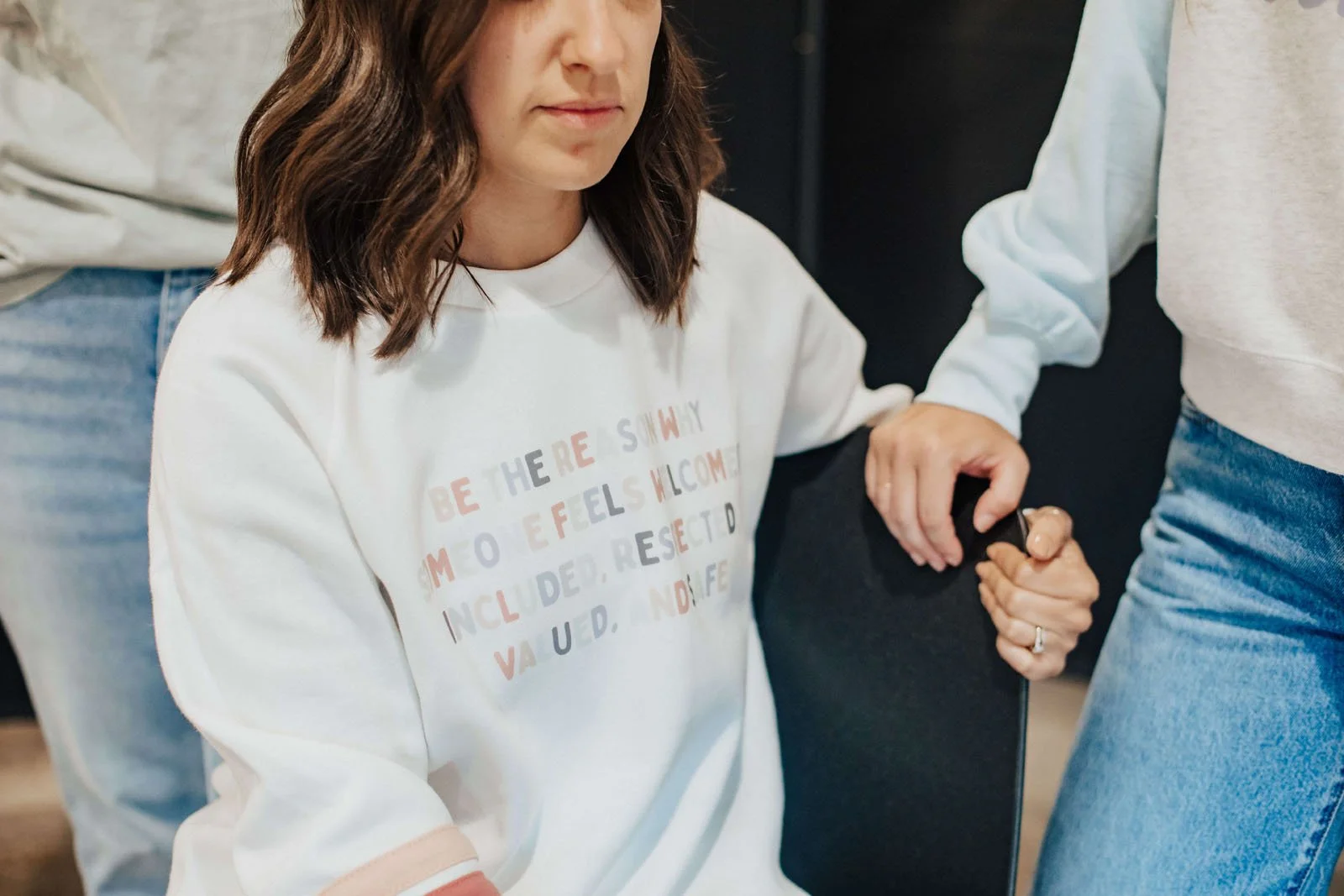 A woman with shoulder-length brown hair sitting with her eyes closed, holding hands with another person while seated. The woman wears a white sweatshirt with colorful text. The other person, wearing a light gray sweatshirt, stands beside her.