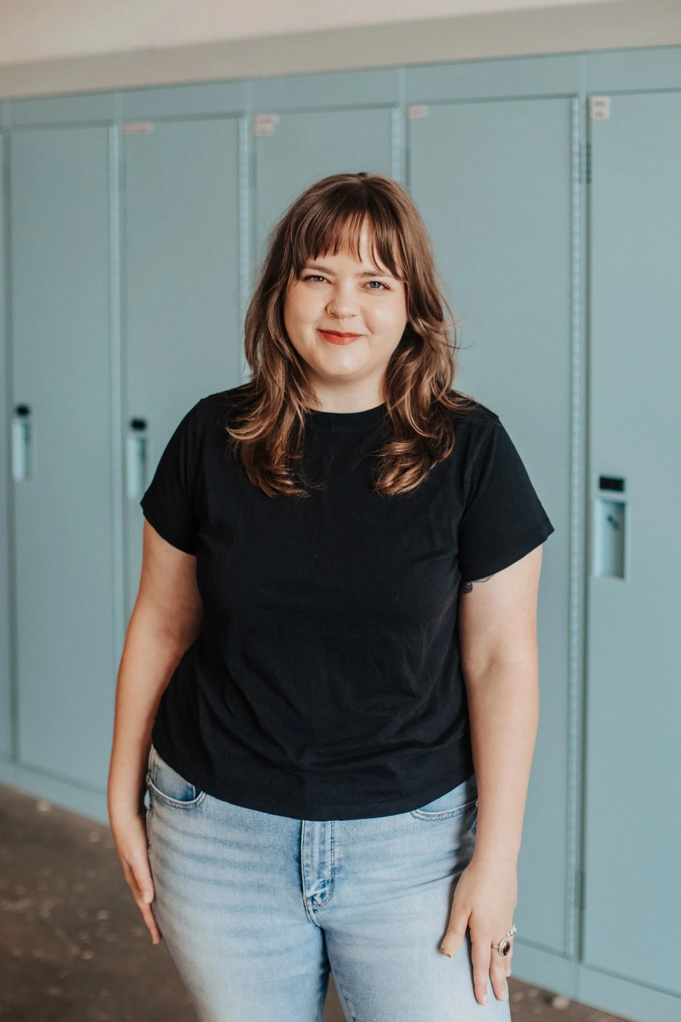 A young woman with shoulder-length brown hair, smiling and wearing a black T-shirt and light blue jeans, standing in front of lockers.