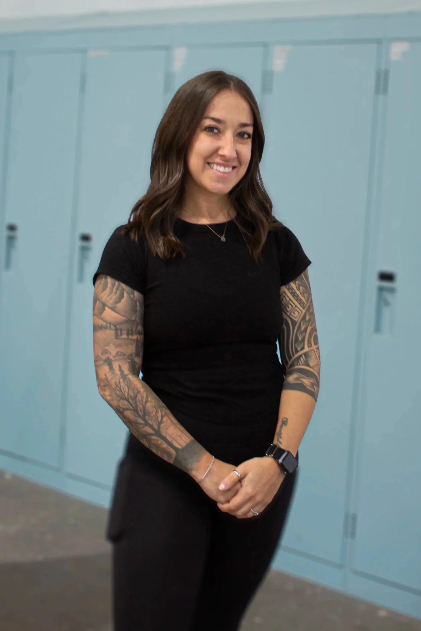 A smiling woman with shoulder-length brown hair, tattoos on both arms, wearing a black t-shirt, blue jeans, and a smartwatch, standing in front of light blue lockers.