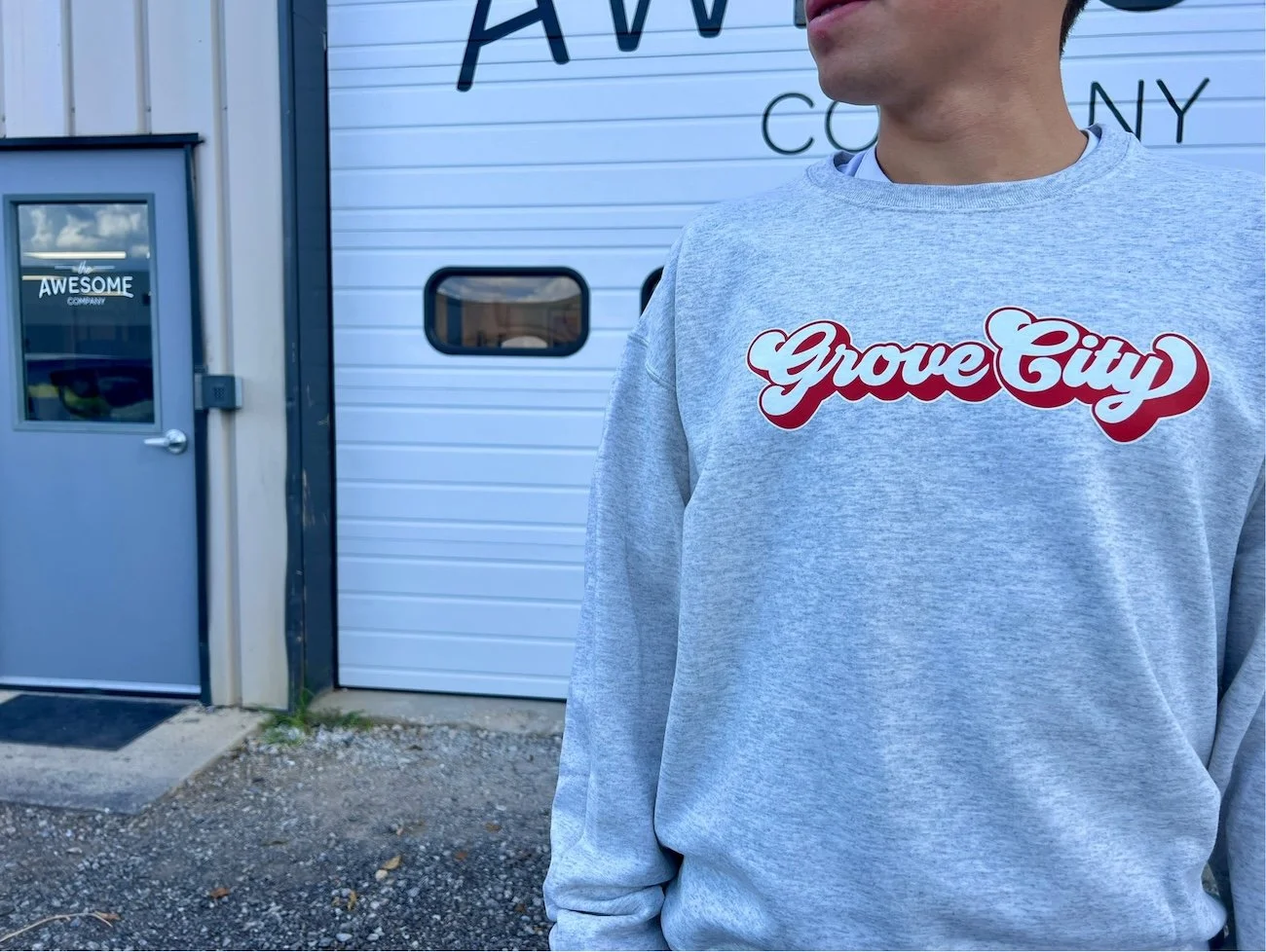 A person wearing a gray sweatshirt with red and white text reading "Grove City" standing outside near a building with a white garage door and a gray door with a window that has a sticker reading "the AWESOME".