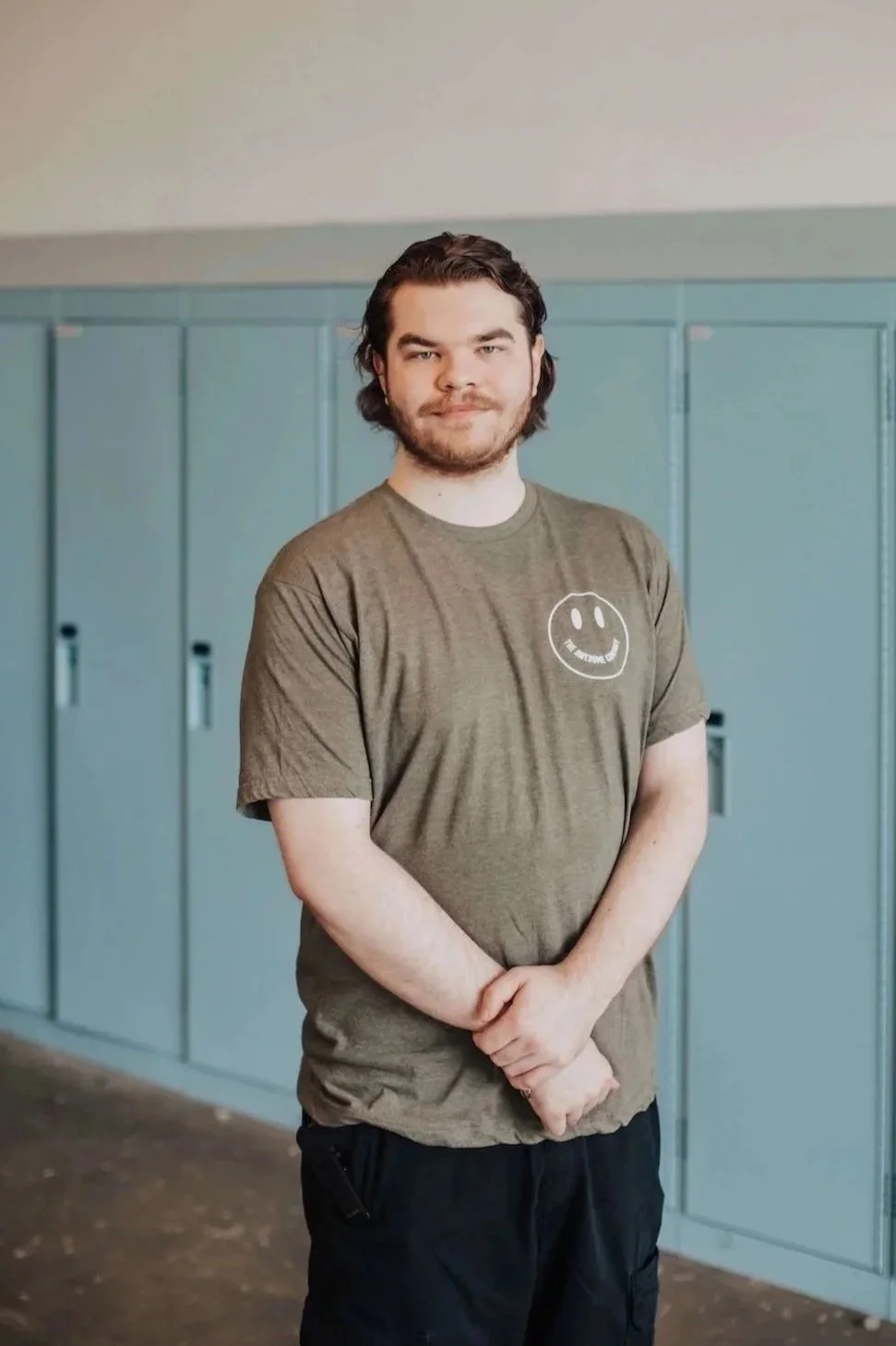 A young man with dark hair, a beard, wearing a green T-shirt with a smiley face logo and black pants, standing in front of light blue lockers in a school hallway.