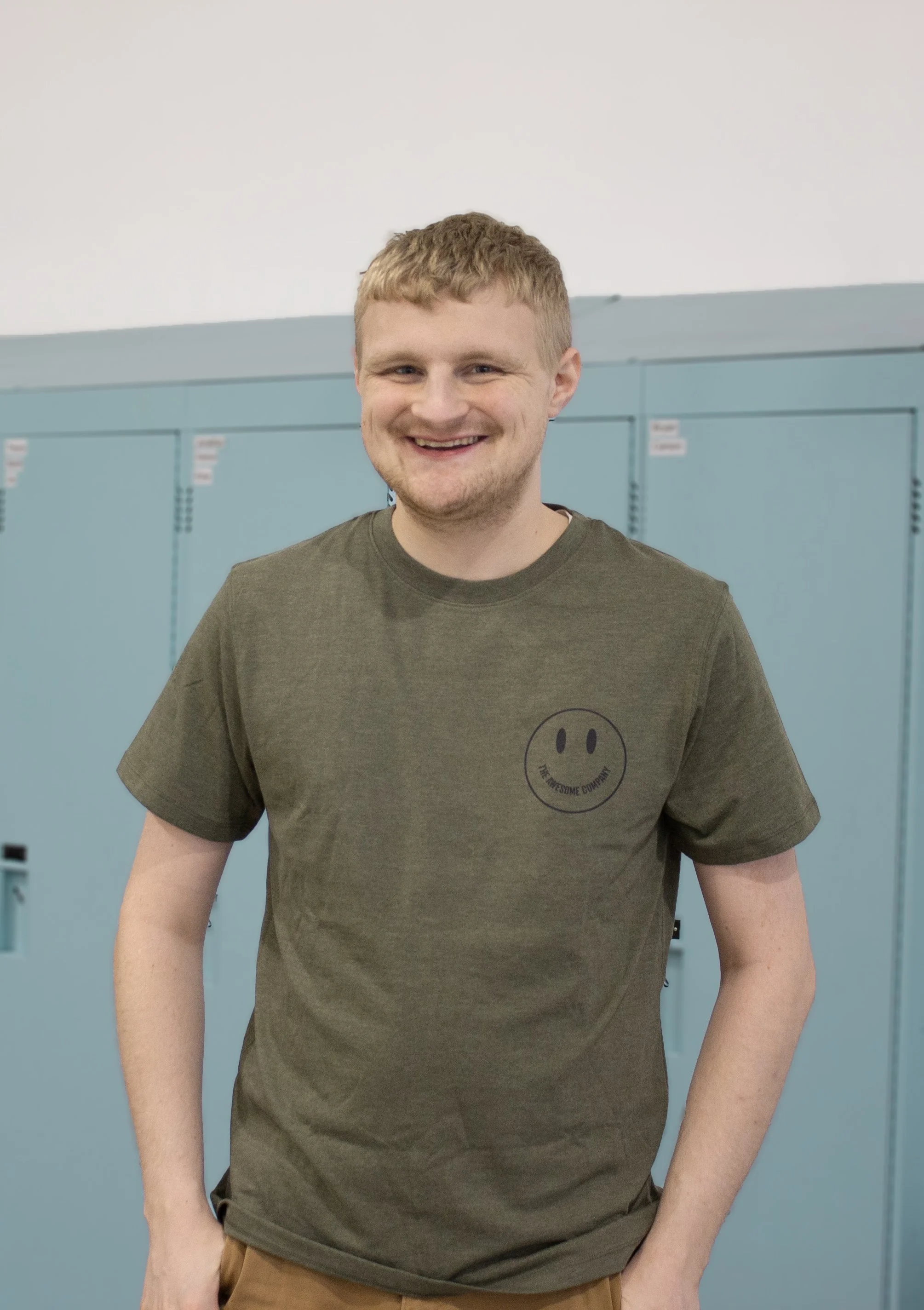 A young man with blond hair and a beard, smiling, wearing a green t-shirt with a smiley face logo, standing in front of blue lockers.