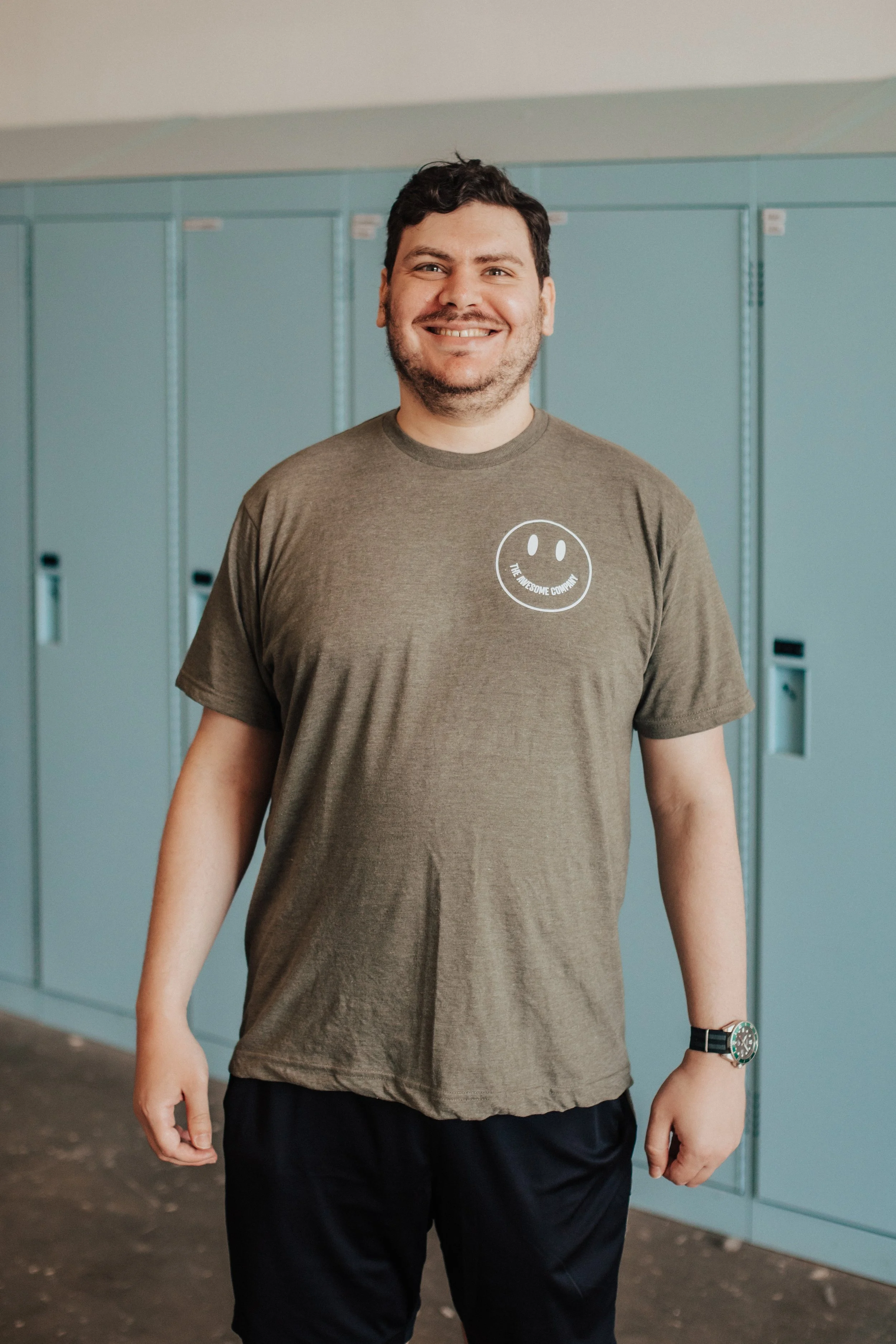 A smiling man with dark, curly hair and a beard, wearing a gray T-shirt with a smiley face design and the text 'The Awesome Company,' standing indoors in front of blue lockers.