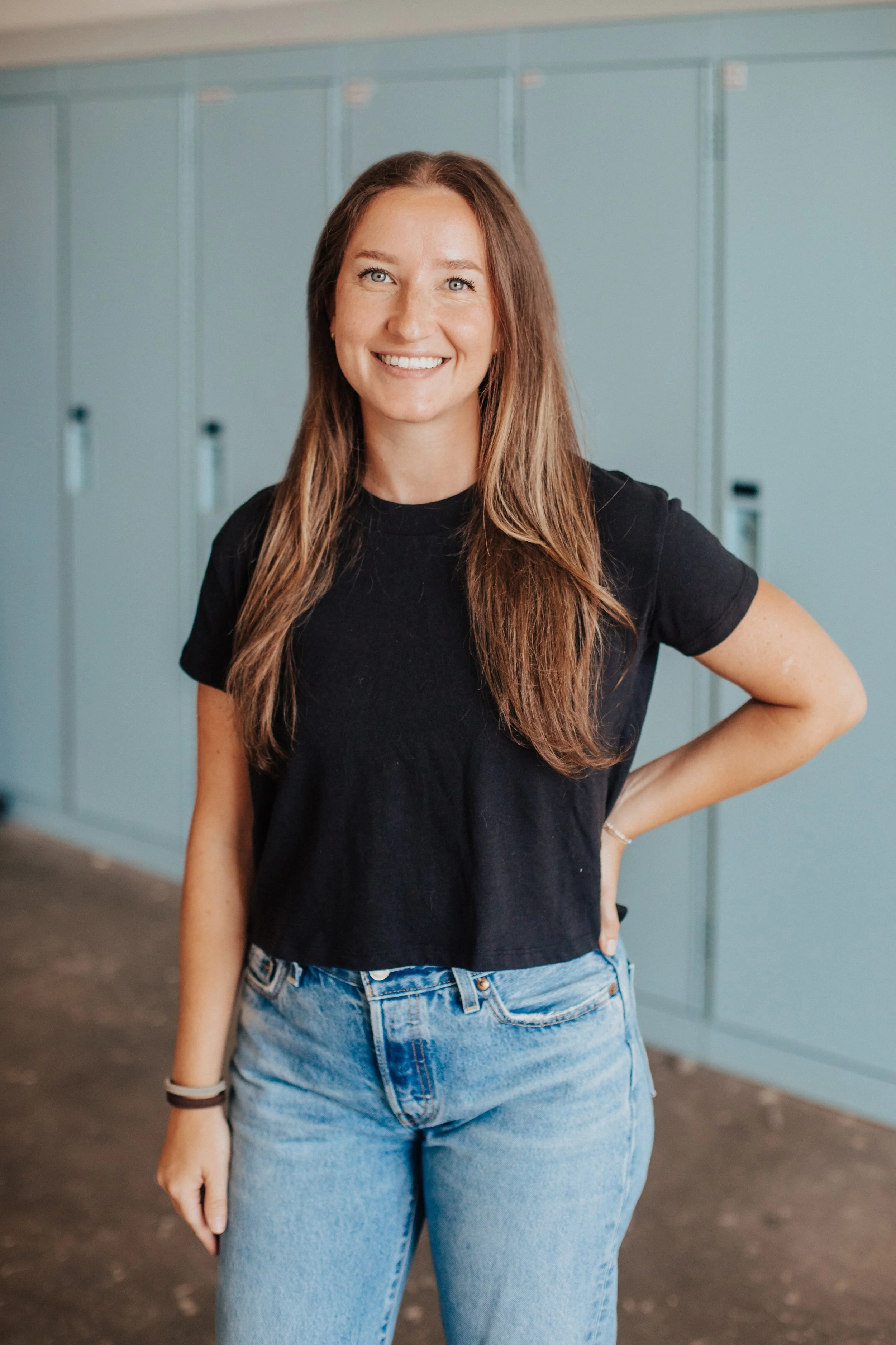 A young woman with long brown hair and blue eyes smiling, wearing a black T-shirt and light blue jeans, standing in front of green lockers.