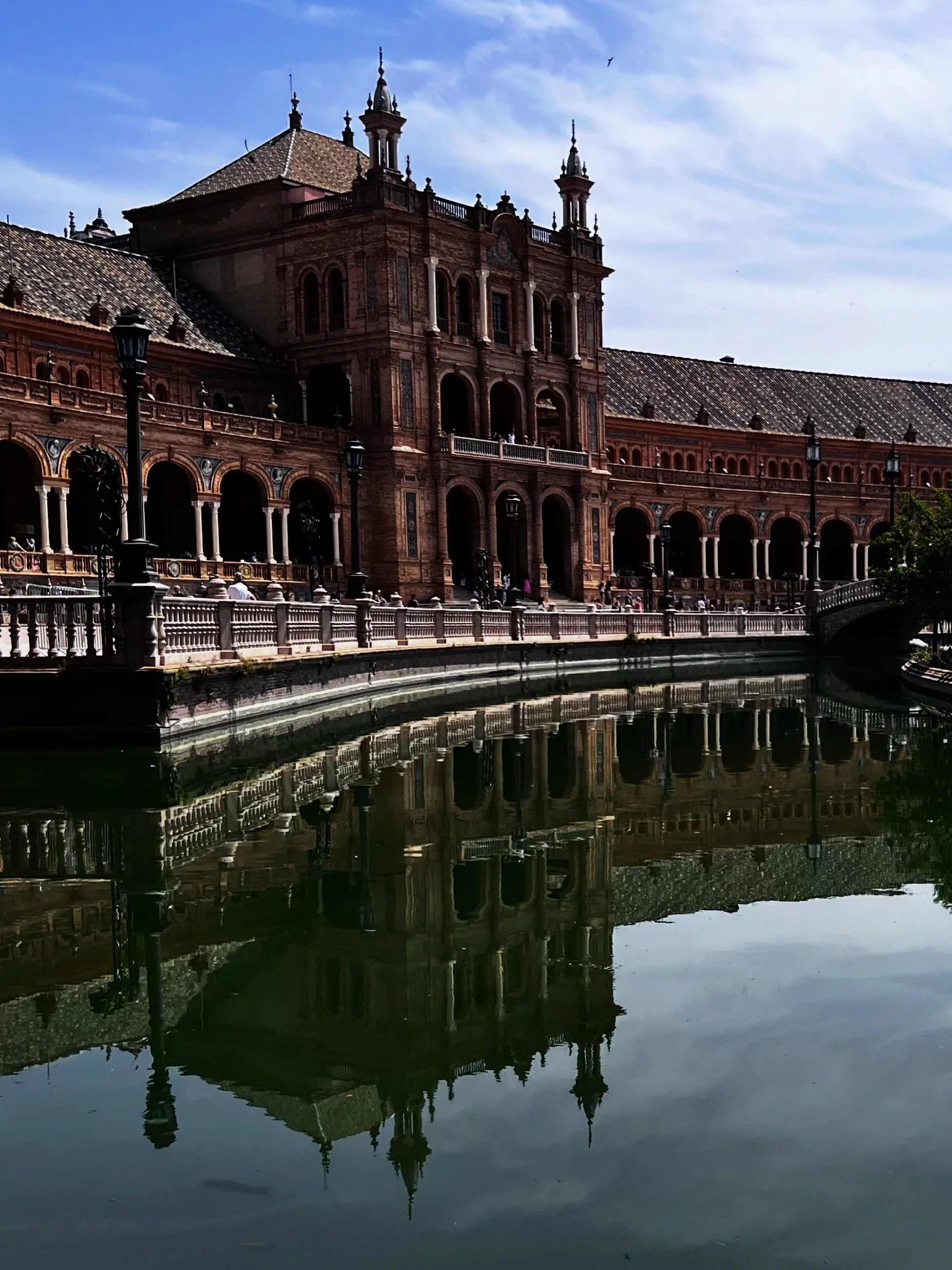 Historic building with ornate architecture, arches, and spires, reflected in a calm water body, under a blue sky with scattered clouds.