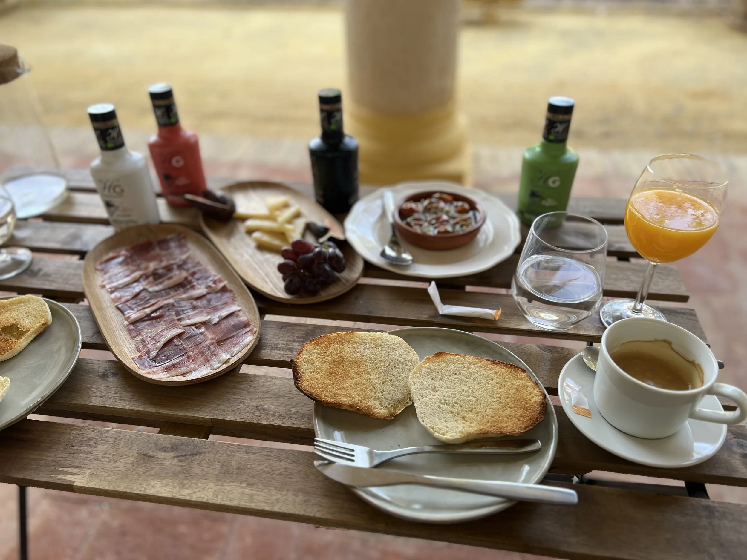 Breakfast spread on a wooden table including toasted bread, sliced cured meat, grapes, cheese, a bowl of soup, a cup of coffee, a glass of orange juice, and bottles of various sauces or condiments.