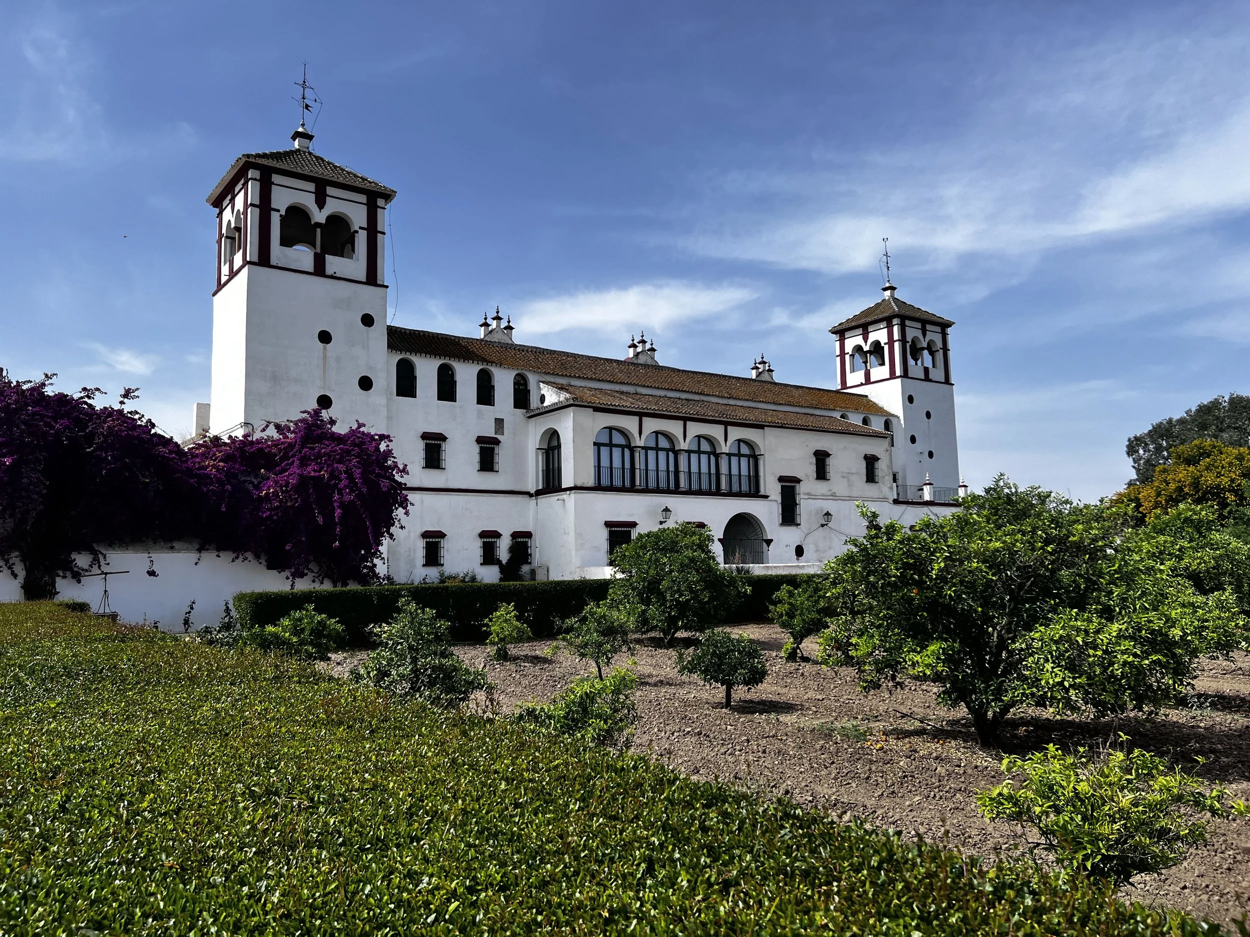 A white Spanish-style mansion with two towers, surrounded by lush green garden and purple flowering bushes, against a blue sky with some clouds.