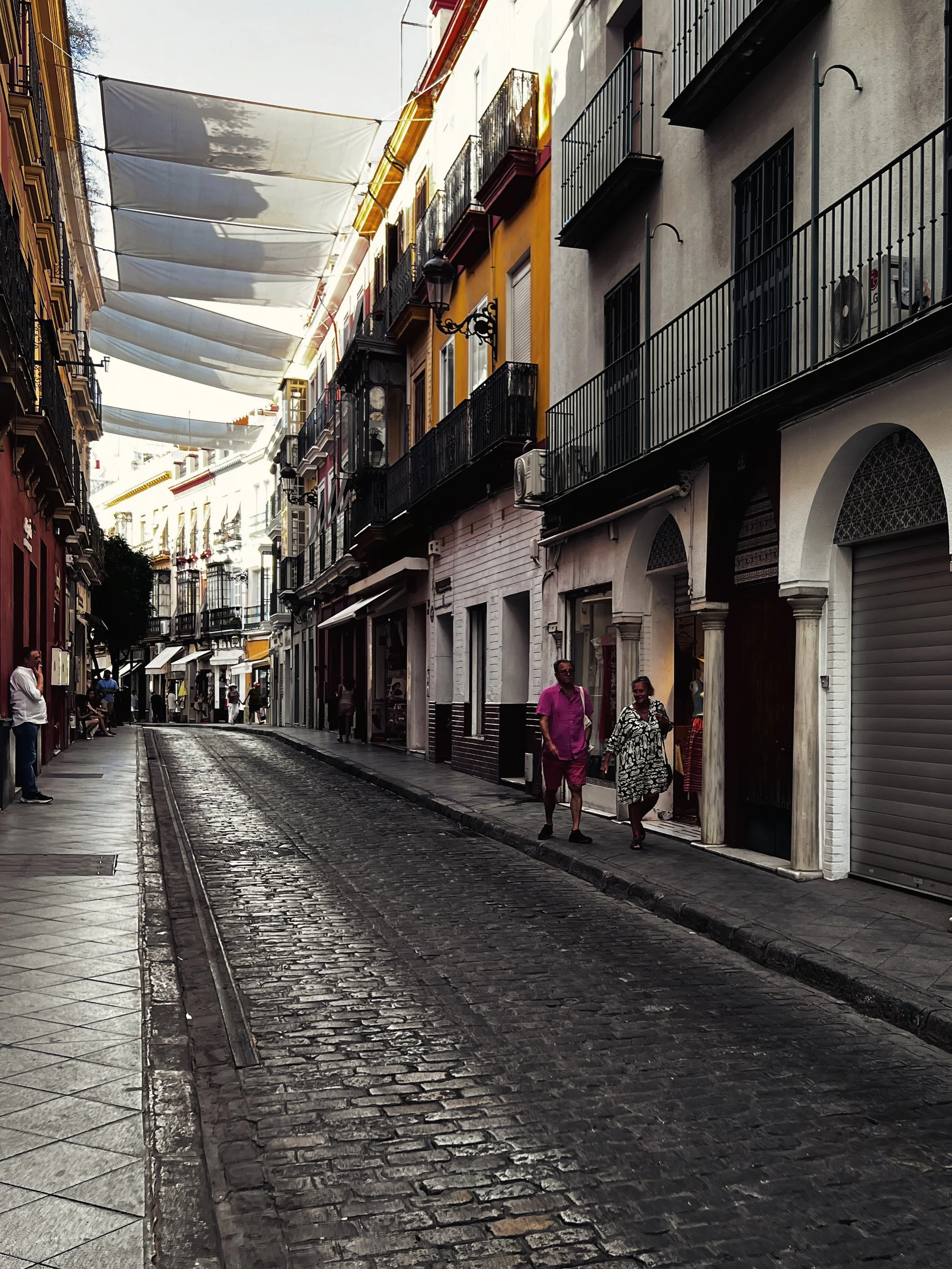 A narrow cobblestone street in a European city with colorful buildings and balconies, some with laundry hanging above. Several pedestrians walk along the sidewalk, and the sky is partly covered with fabric or shade cloths.