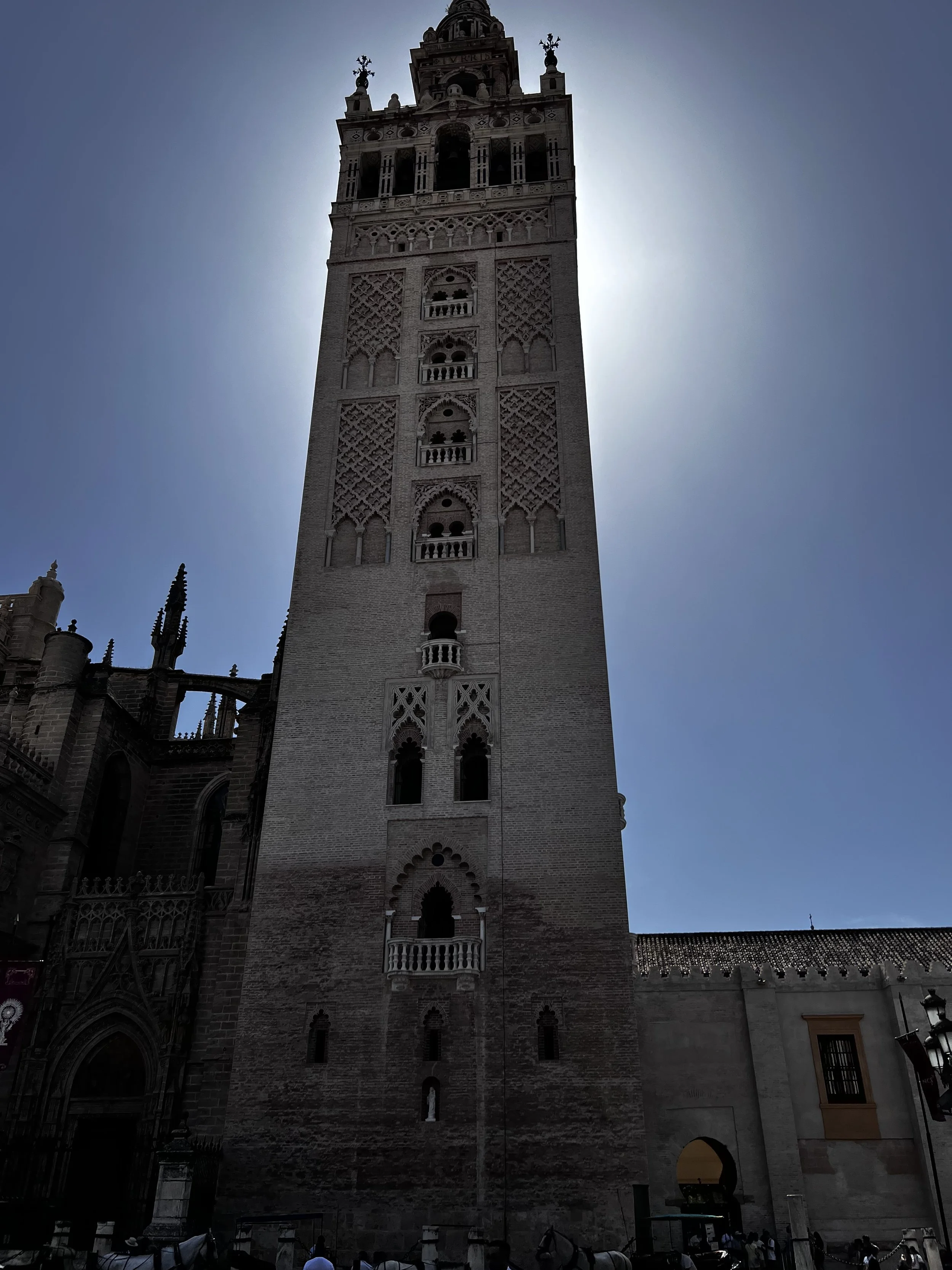 The image shows a tall, Gothic-style bell tower with detailed architecture silhouetted against a bright sun and clear blue sky.