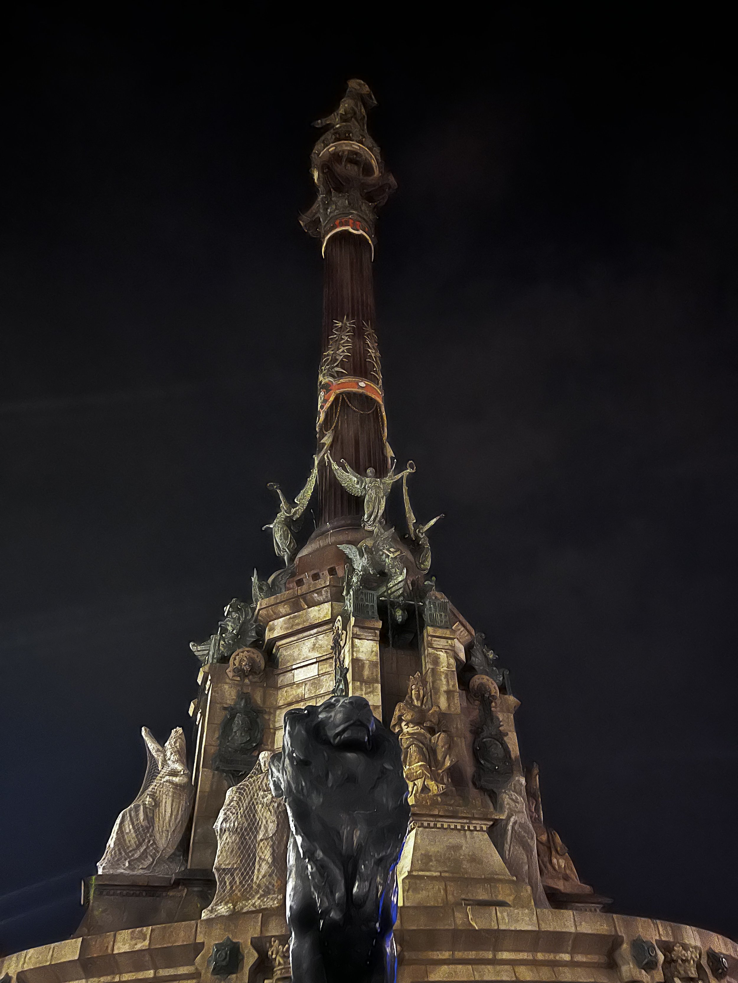 Nighttime view of the Columbus Monument in Barcelona, Spain, featuring a tall column topped with a statue of Christopher Columbus, with decorative sculptures and statues at the base, illuminated against the dark sky.