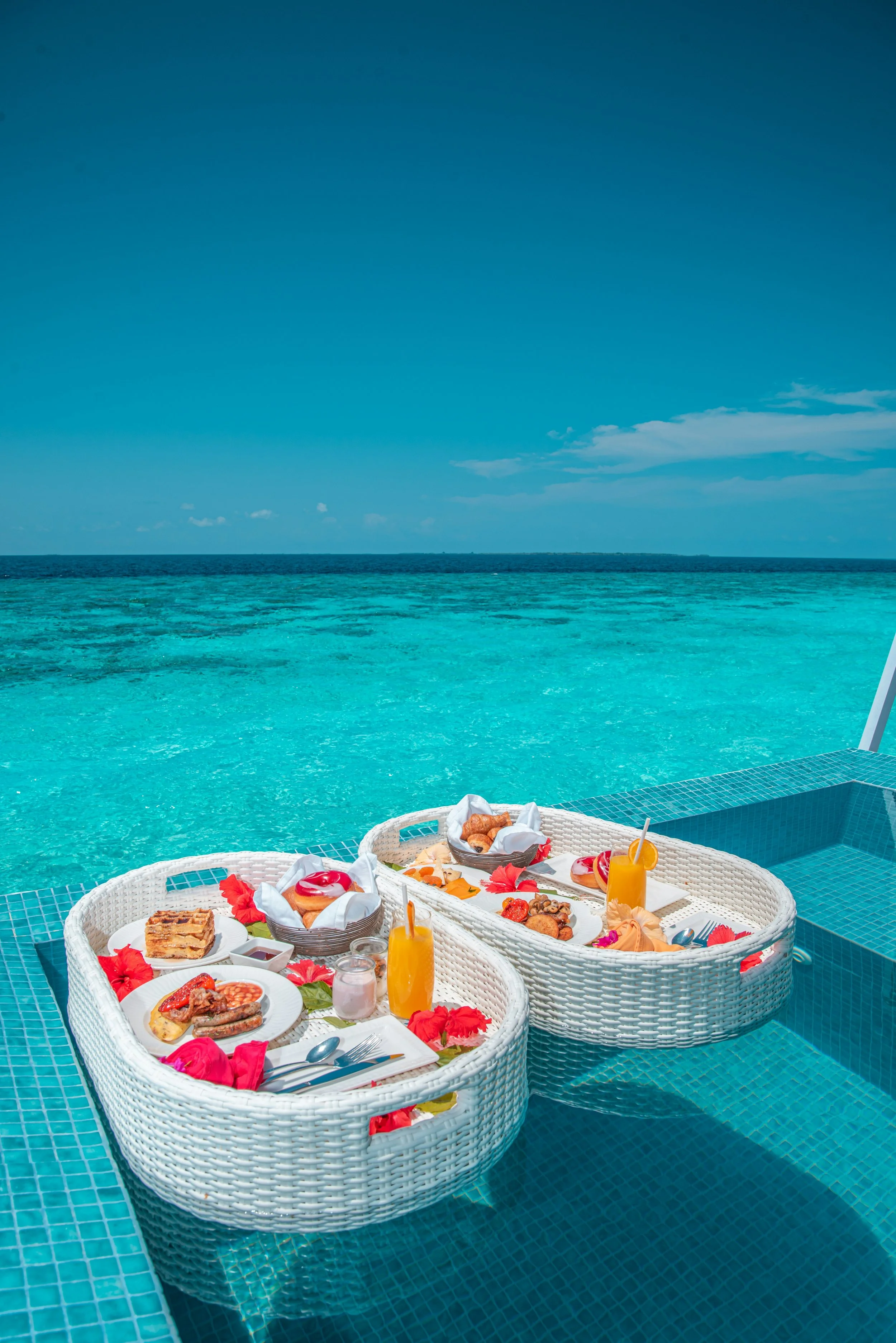 A poolside setup with white woven baskets filled with breakfast foods and drinks, floating on a blue-tiled pool edge overlooking the ocean under a clear blue sky.
