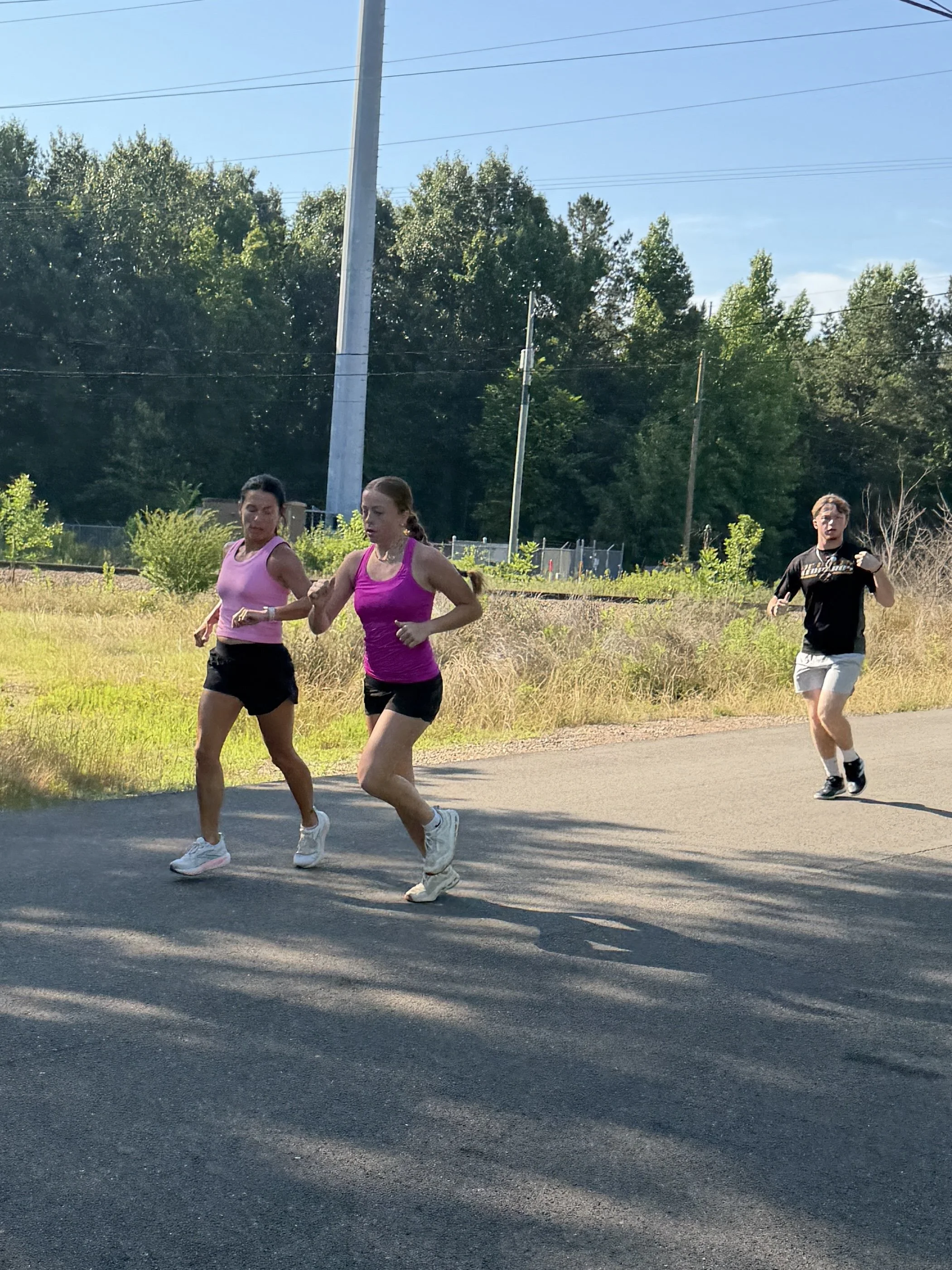 Three people, two women and one man, running on a paved path in a grassy area with trees and utility poles in the background on a sunny day.