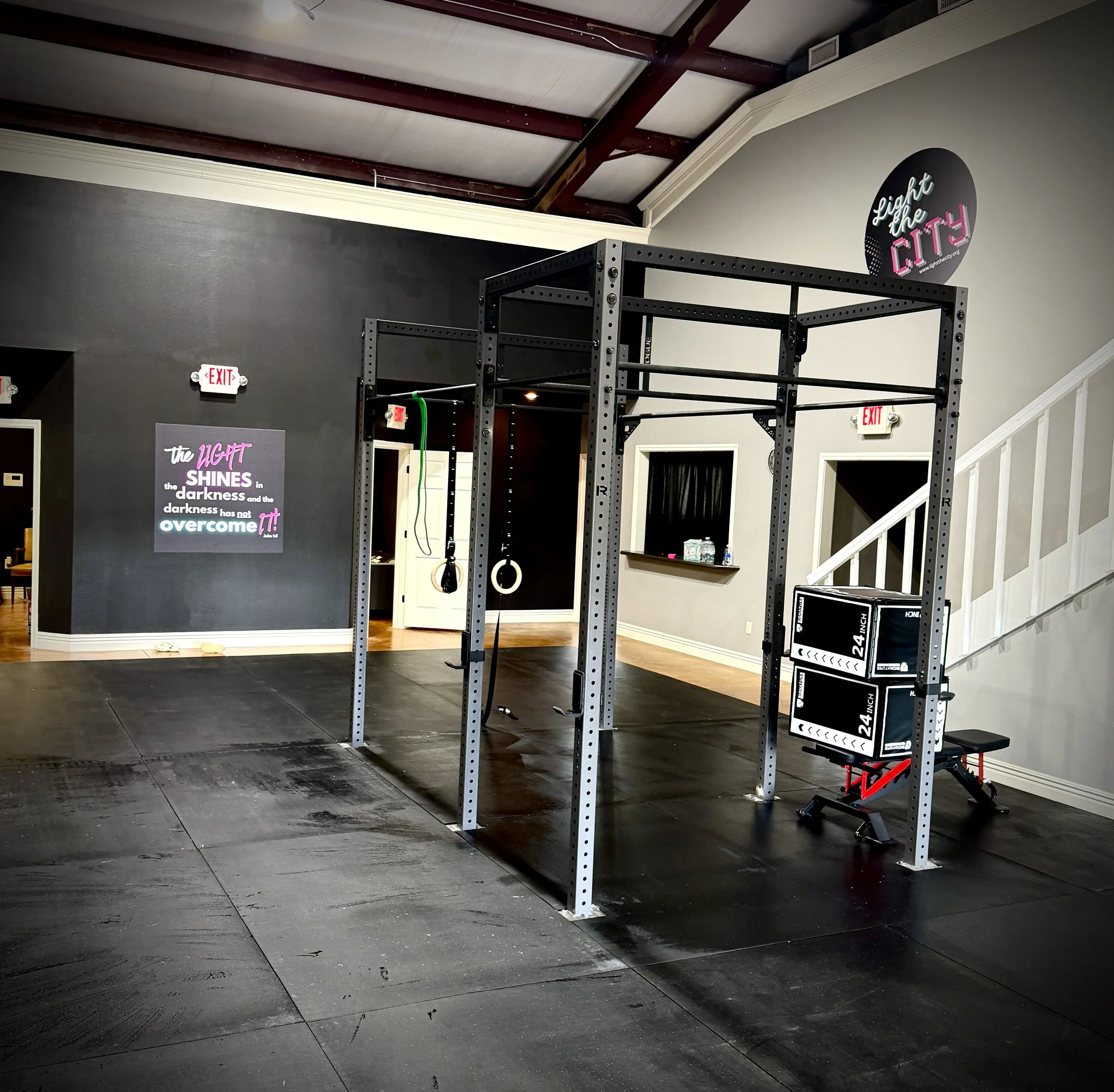 Interior of a gym or fitness center with a squat rack, gymnastic rings, and motivational posters on the wall. The gym has dark flooring and a wall with signs and quotes, including one that says "the LIGHT SHINES in the darkness and the darkness has not over come it!"